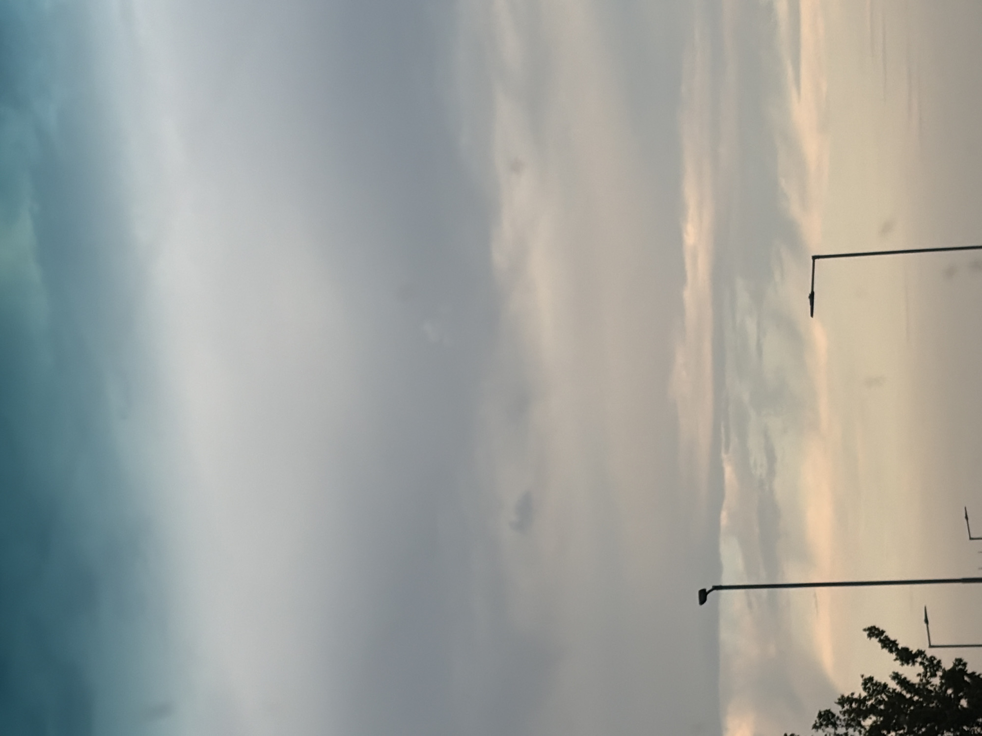 The image shows a cloudy sky with a hint of teal and pink hues, suggesting either dawn or dusk. In the foreground, silhouetted streetlights stand tall against the sky. One streetlight is prominently positioned in the lower left quadrant, and another is visible to its right. Further to the right, a third streetlight is partially visible near the bottom edge of the frame. A portion of a tree with dark leaves is visible in the far left, adding a natural element to the scene. The overall atmosphere is serene and somewhat muted, typical of an overcast sky. The scene is not specific to Nitra, Slovakia, or any identifiable city, as it lacks distinctive landmarks or urban features.