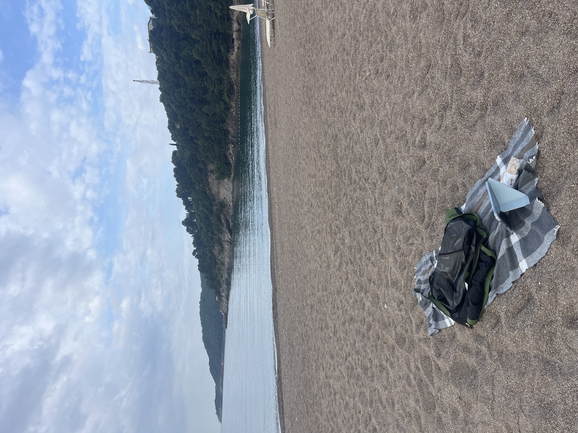 The scene is a beach at Boreti, Montenegro. The sky is partly cloudy. A mountain covered in trees is to the left of the frame. The Adriatic Sea is in the middle of the frame. A grey sand beach occupies the right side of the image. On the beach is a grey and white plaid blanket, a black and green backpack, a white triangular object (possibly a book), and a plastic bag of food.