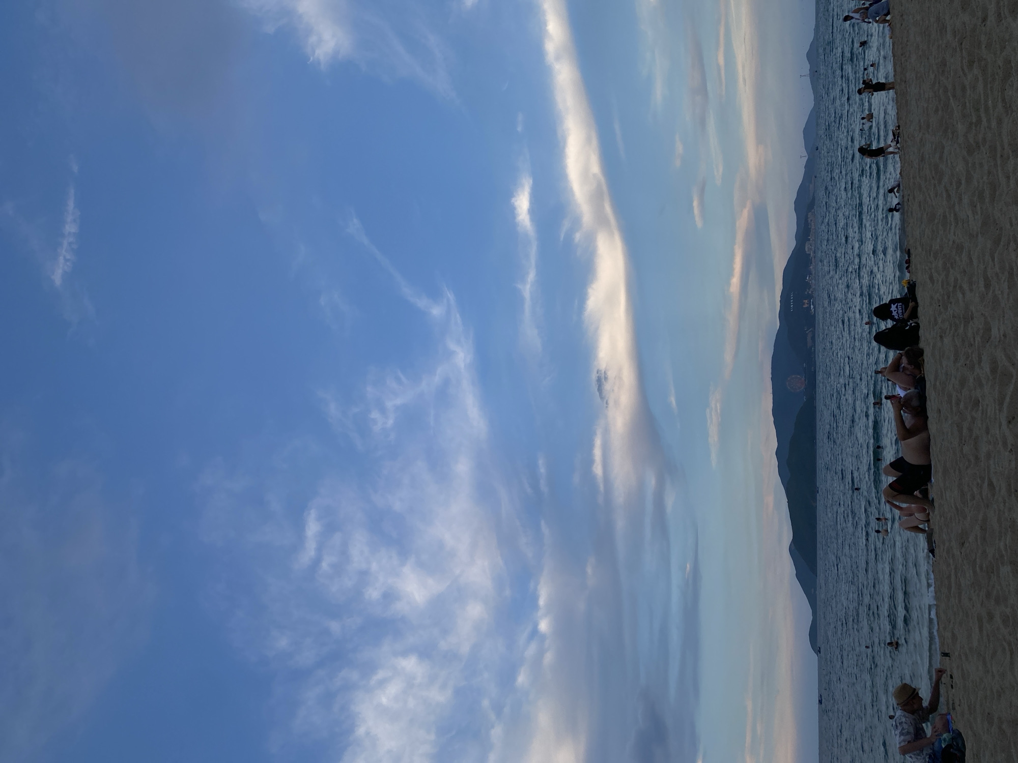 The image captures a serene beach scene in Nha Trang Ward, Vietnam, during what appears to be the late afternoon or early evening. The sky is a canvas of blue, adorned with wispy white clouds that are beginning to take on hues of pink and orange, hinting at an approaching sunset. In the distance, a lush, green mountain range forms a dramatic backdrop to the city, with buildings and a Ferris wheel visible on its slopes.
The foreground is dominated by a sandy beach, where several individuals are relaxing and enjoying the tranquil atmosphere. A group of people are lying on the sand, some facing the sea, others conversing. Closer to the water's edge, a man wearing a hat is interacting with objects on the sand, possibly playing a game or examining something.
The sea is a deep blue, with gentle waves lapping at the shore. Several people are visible in the water, swimming or wading. Boats are scattered across the horizon, adding to the sense of a thriving coastal community. The overall impression is one of a peaceful and idyllic setting, perfect for unwinding and enjoying the natural beauty of Nha Trang.