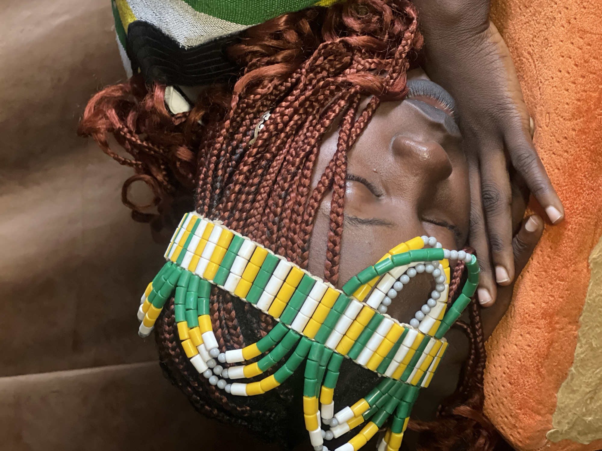 The image depicts a person sleeping on their side, with their head resting on their arms. They have reddish-brown braids and are wearing a colorful beaded headband with green, yellow, and white cylindrical beads. The person is lying on an orange textured surface, possibly a blanket or cushion. In the background, there is a brown textured surface, which could be upholstery or fabric. To the right of the person's head, a portion of a patterned textile with green, white, and black stripes is visible. The scene appears to be indoors, possibly in a domestic setting. There is no indication of the specific location within Jos, Nigeria.