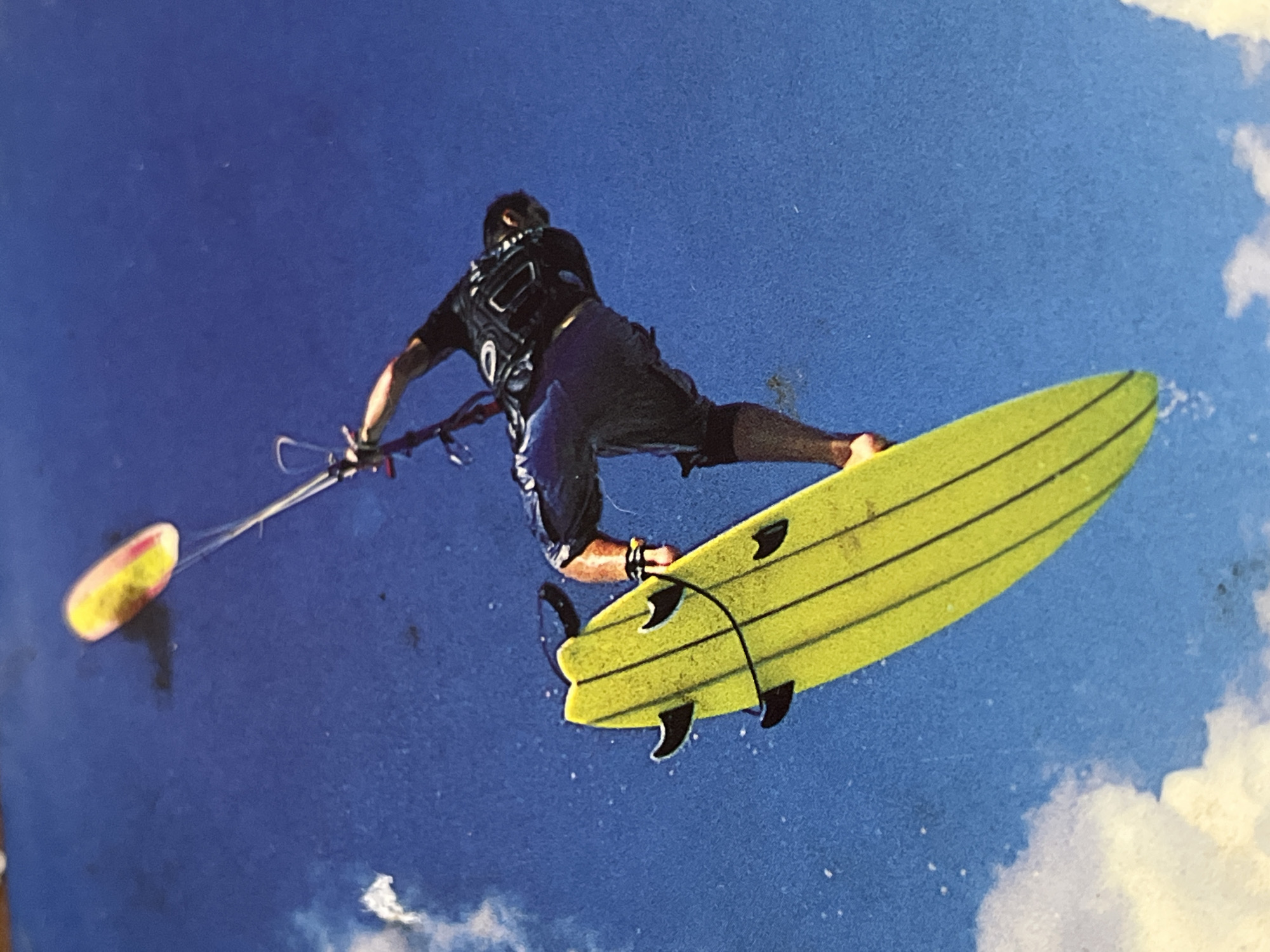 The image depicts a kitesurfer in action against a partly cloudy blue sky. The kitesurfer, seen from the back, is wearing a black top and dark-colored shorts. They are holding a kite control bar, with the kite itself appearing as a yellow-and-red object. The kitesurfer is airborne, riding a yellow surfboard with black fins. White clouds are visible in the lower right corner. The location is not specified and cannot be determined from the image.