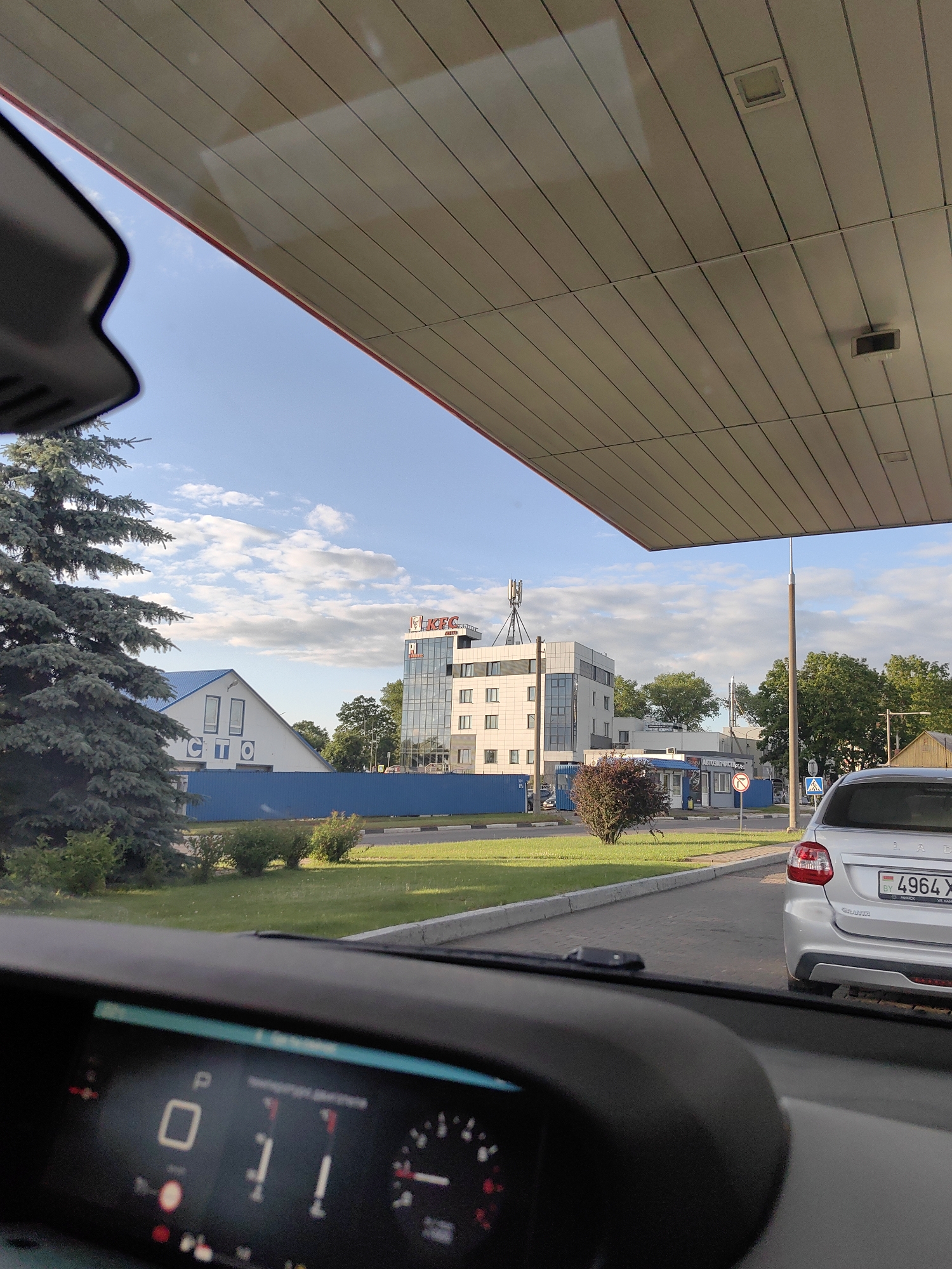 An exterior view from inside a car, looking out over a gas station canopy. The car's dashboard is partially visible in the foreground. In the middle ground, across a paved area, there is a building with a blue pitched roof and white walls, with the Cyrillic letters "СТО" visible on its facade. To the right of this building, a modern, multi-story white building with glass windows is visible, featuring a red KFC logo at its top. A silver LADA car with a Belarusian license plate is parked to the right of the frame. The environment is an urban or suburban setting with green grass, trees, and a blue sky with scattered clouds. The time appears to be daytime, likely late afternoon or early evening due to the lighting. The location is identified as the city of Фаніпальскі сельскі Савет, Belarus.