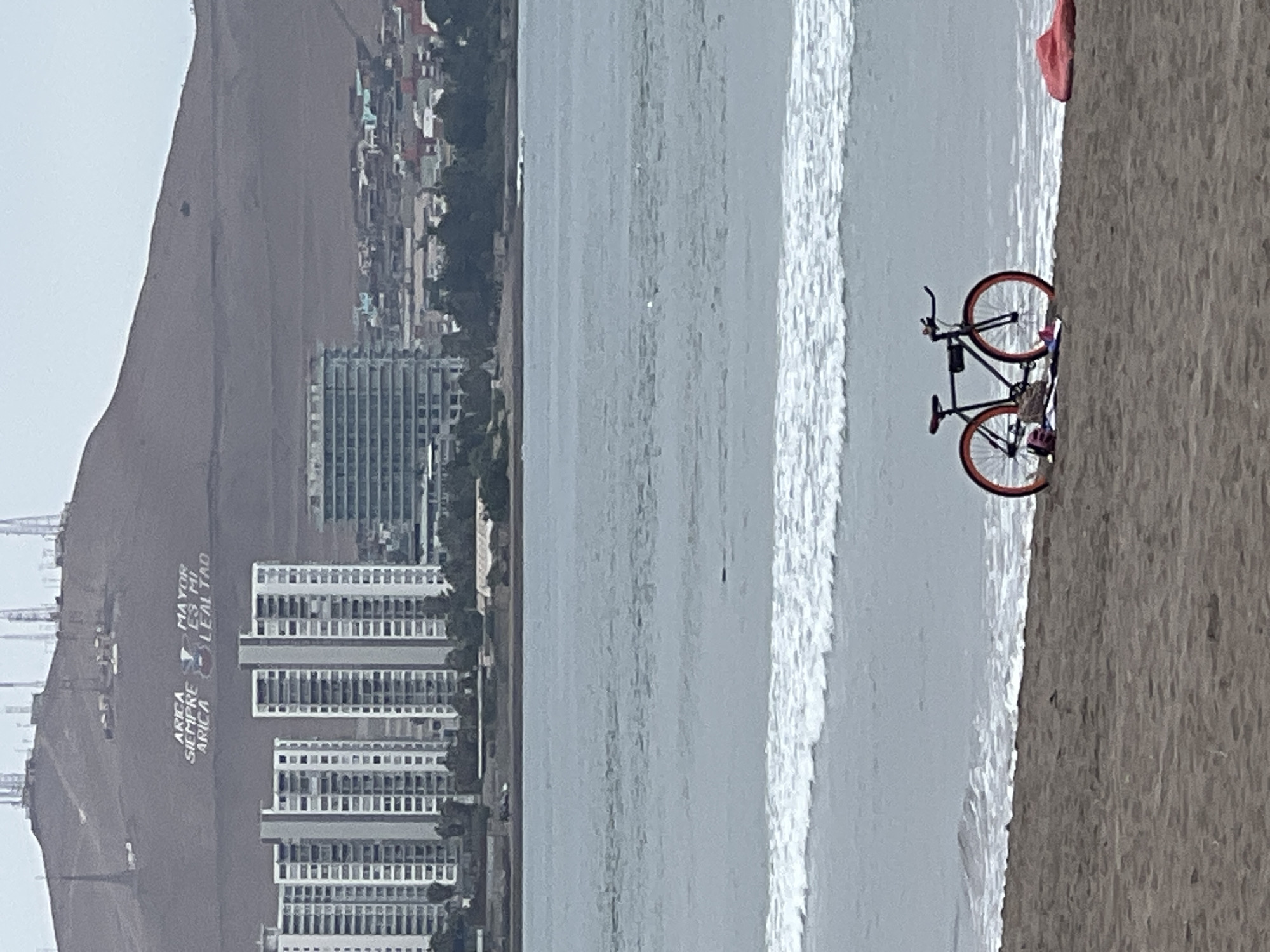A vertical, wide shot depicts a beach scene in Arica, Chile. The beach runs along the right side, marked by a sandy terrain. In the middle, ocean waves crash onto the shore. To the left of the ocean, the city of Arica is visible, with multiple buildings clustered together. Closer to the foreground, tall residential buildings stand out. Further back, on a hill overlooking the city, white lettering reads "ARICA SIEMPRE ES FIEL" and "ARICA LA LEALTAD."
On the right side of the beach, a bicycle is parked, leaning against the sand. The bike has black frames and red tires. Adjacent to the bicycle, a pink object is seen on the sand.