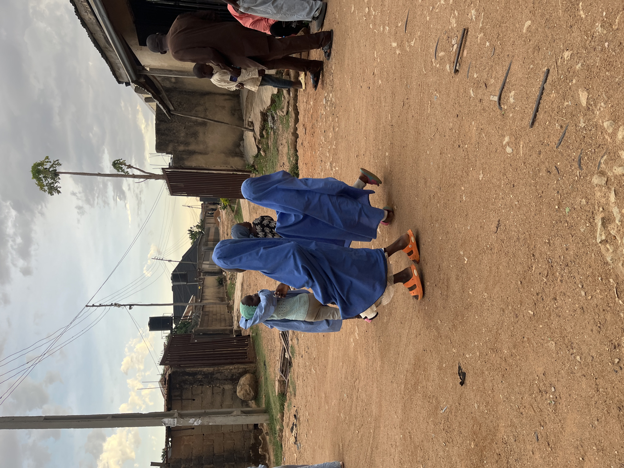 Two individuals wearing long blue robes and orange sandals walk along a dirt road. Buildings constructed of concrete blocks and corrugated metal line the street. Overhead power lines traverse the scene. In the distance, a tall tree stands beside a building. The location is Barakin Akawo, Nigeria.