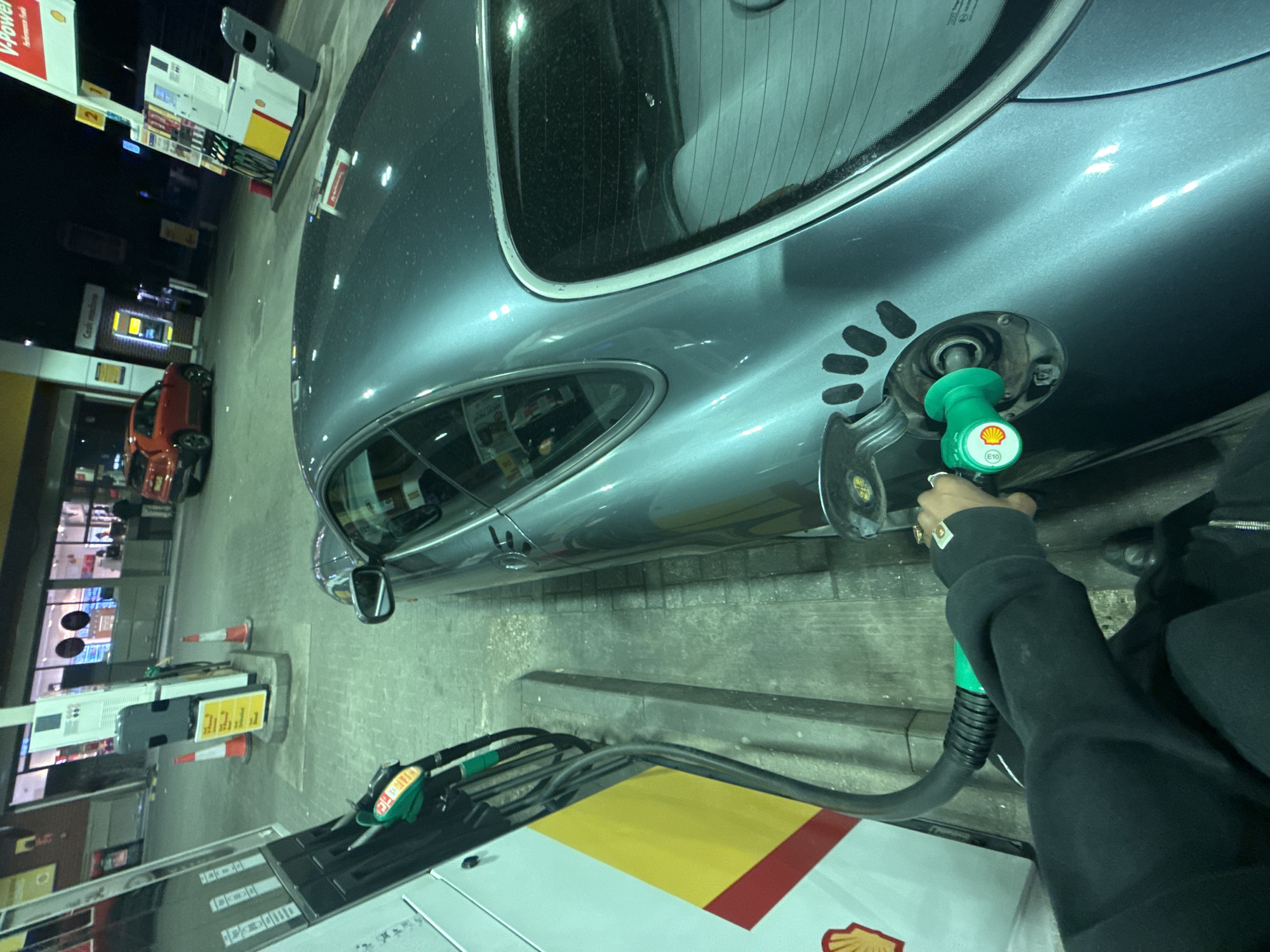 The image captures a nighttime scene at a Shell gas station, likely in London, United Kingdom. The primary focus is a grey car, possibly a Porsche, being refueled. A person's arm and hand, wearing a dark long-sleeved top and rings, are visible holding a green Shell-branded fuel nozzle. The nozzle is inserted into the car's fuel tank opening, which is surrounded by a pattern of five small black oval shapes on the car's body. The nozzle clearly displays "E10", indicating the type of fuel being dispensed.
In the background, the gas station's infrastructure is illuminated, contrasting with the dark night sky. Several white and yellow Shell fuel pumps are visible, with one displaying "V-Power" on its sign. Another sign in the distance reads "Cash machine." A blurred red car is parked further away at another pump. Orange road cones are placed near one of the pumps. The ground appears to be concrete or paved. The car itself has a sleek, metallic grey finish, with noticeable dust or specks on its surface, particularly on the rear window. The overall ambiance is functional and typical of a late-night gas station stop.