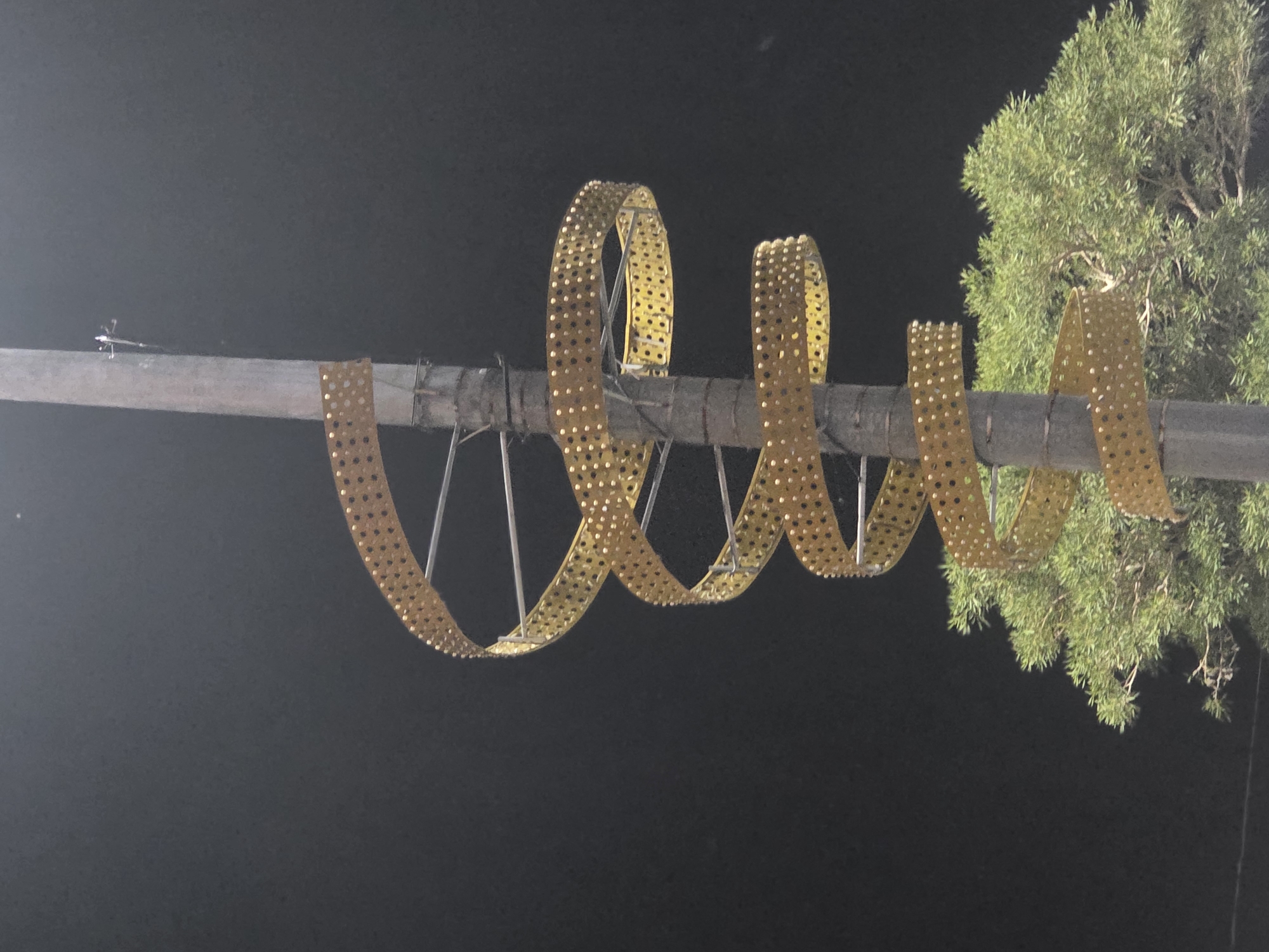 The image shows a tall, grey, cylindrical pole with a spiral structure wrapped around it. This spiral structure is made of a golden, perforated material and is supported by thin metal rods. The spiral descends down the pole. At the base of the pole, a lush green tree is visible. The background is a dark, uniform sky, suggesting the photo was taken at night. The scene is located in Padang Aro, Indonesia.