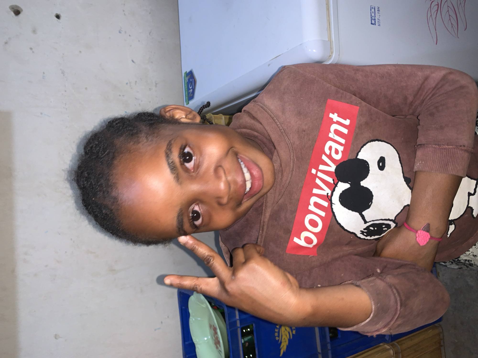 A young girl with dark skin and braided hair is centered, smiling broadly with an open mouth and dark brown eyes, posing for the camera. She is wearing a reddish-brown long-sleeved hoodie featuring a red rectangular graphic with white text "bonvivant" and a large white cartoon character wearing sunglasses below it. Her right hand is raised, forming a peace sign (V-sign) with her index and middle fingers extended. On her left wrist, she wears a thin pink bracelet with a pink heart charm. The indoor background consists of a light-colored, stained concrete wall. To the left, stacked blue plastic crates are visible, with a light green plastic plate on top of the uppermost crate. To the right, a white upright refrigerator or freezer with "No. 1" and "HTF-19H" labels is present. This scene is located in New Stadium, Nigeria.