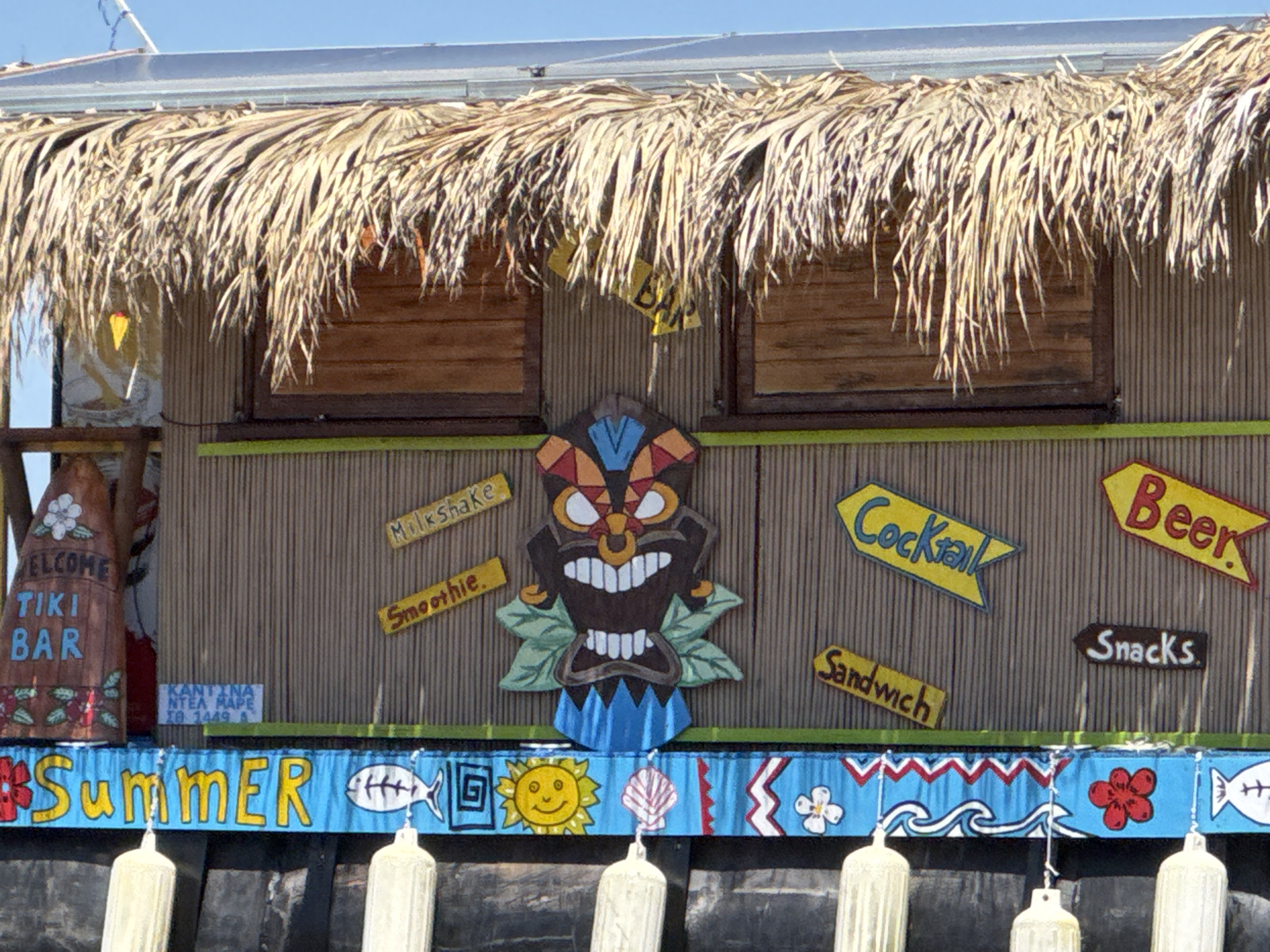 The image displays the front of a colorful, outdoor tiki bar under a clear blue sky. The structure features a distinctive straw or thatch roof and walls made to resemble vertical bamboo reeds. Two closed wooden-slatted windows are visible on the facade.
A large, stylized tiki mask decoration, painted in shades of brown, red, blue, and yellow with a grinning mouth, is centrally positioned on the wall. Surrounding the tiki mask are several arrow-shaped wooden signs advertising the bar's offerings: "Milkshake" and "Smoothie" on the left, and "Cocktail," "Beer," "Sandwich," and "Snacks" on the right.
To the far left, a tall, brown sign with floral accents proclaims "WELCOME TIKI BAR" in blue lettering. Below this, a small white sign with blue text reads "KANTINA NTEA MAPA ΣΑ 1449 Α," providing a localized context.
A decorative blue banner runs along the bottom of the facade, prominently displaying the word "Summer" in yellow letters. The banner is adorned with various maritime and summery motifs, including white fish, a yellow smiling sun, a white seashell, blue and white waves, and red and white flowers. Hanging below this banner are several white cylindrical objects, possibly boat buoys, suggesting the bar is located near or on water.
The scene is brightly lit by natural sunlight, indicating daytime. The vibrant decorations and offerings suggest a casual, recreational setting, likely a beach or waterfront area in the Sithonia Municipal Unit, Greece, as hinted by the Greek text on the small sign. No people are visible.
