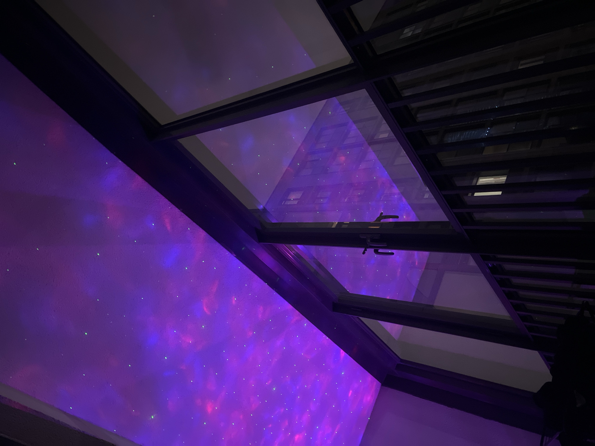 The image shows a view through a window towards the ceiling. The ceiling is illuminated with a projection of a nebula in shades of purple and pink, with small, scattered green points of light resembling stars. The window is divided into several panes with dark frames. Outside the window, through the panes, a glimpse of what appears to be another building can be seen. The precise location in Saint Petersburg, Russia is not discernible from the provided image.