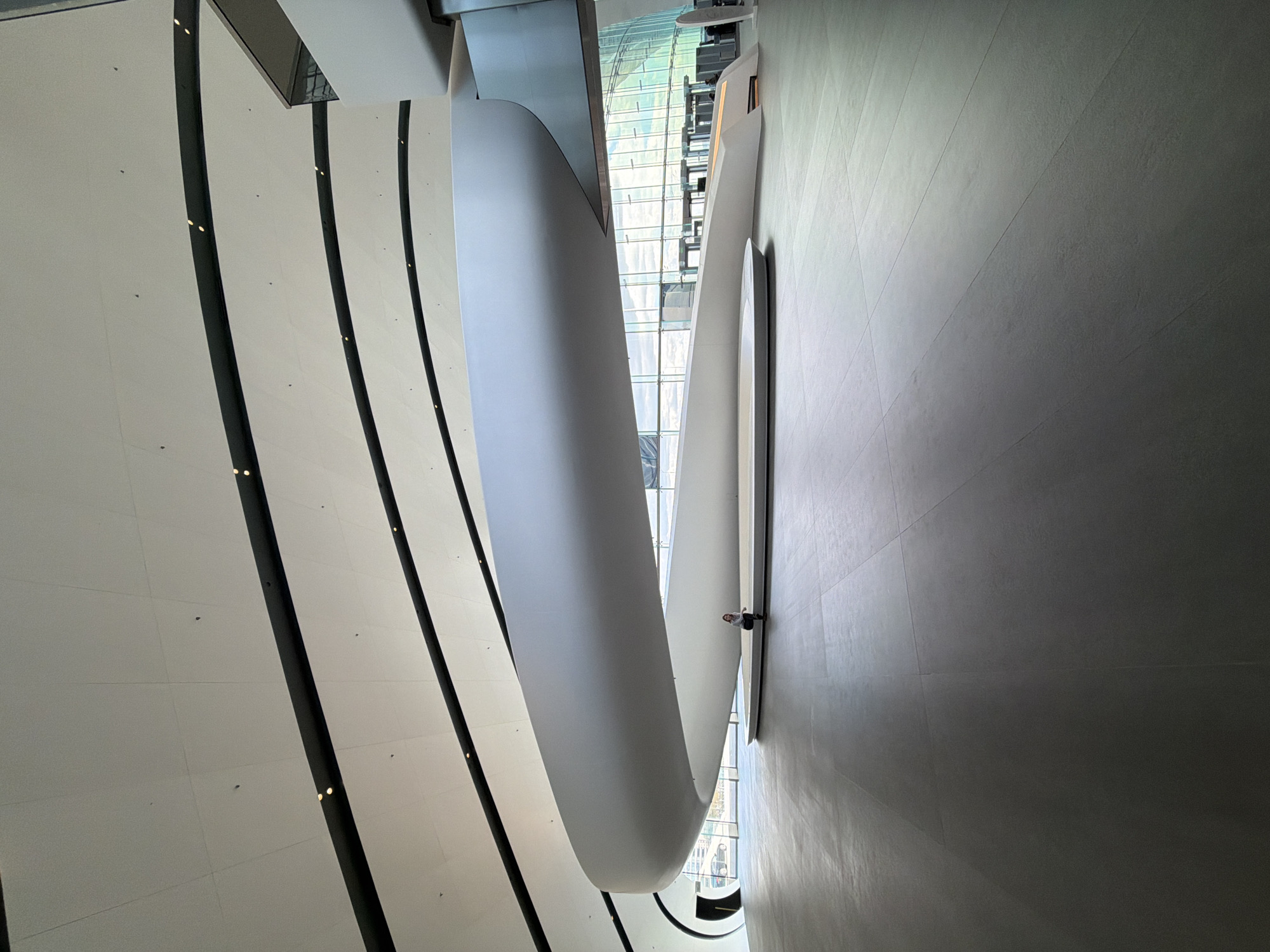 This image depicts the interior of the Lakhta Center in Saint Petersburg, Russia. The perspective is from a high vantage point, looking down into the building's atrium. The architectural design features a spiraling, curved pathway in white, flanked by textured gray walls. The left side of the view shows a wall with regularly spaced vertical black lines and small lights. Visible through the building's glass facade is the city beyond. A single person stands on the curved pathway, facing the gray wall.