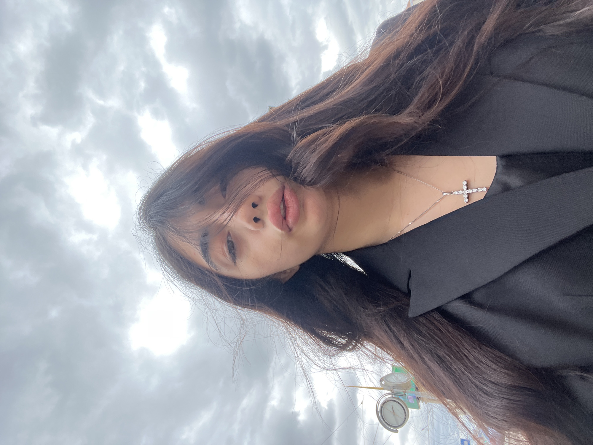 A young woman is taking a selfie outdoors under a cloudy sky, appearing to be in the city of Pilsen, Czechia. She has long, dark, wavy hair that partially covers her face. She is wearing a black blazer over a dark top and a silver necklace with a cross pendant. In the background, there are some indistinct street elements, including what appears to be a clock on a pole. The sky is filled with heavy, gray clouds.