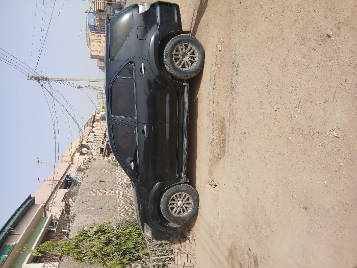 This image displays a black Toyota Fortuner SUV parked on an unpaved, light brown dirt road in Mgbidi, Nigeria. The vehicle is presented in profile, oriented towards the right, and features darkly tinted windows on all side doors and the rear cargo section. The tires show moderate accumulation of dust and dirt, matching the road surface. To the left of the SUV, a slender green, coniferous-type tree grows beside a low brick wall exhibiting a patterned facade, surmounted by security wire. Beyond this wall, a multi-story building with a light green roof is partially visible. The background consists of a street scene lined with various light-colored, multi-story buildings. Numerous overhead utility lines are visible, spanning across a clear blue sky and supported by several utility poles. The dirt road surface is uneven, with scattered small stones and debris. No individuals or active interactions are discernible within the frame.