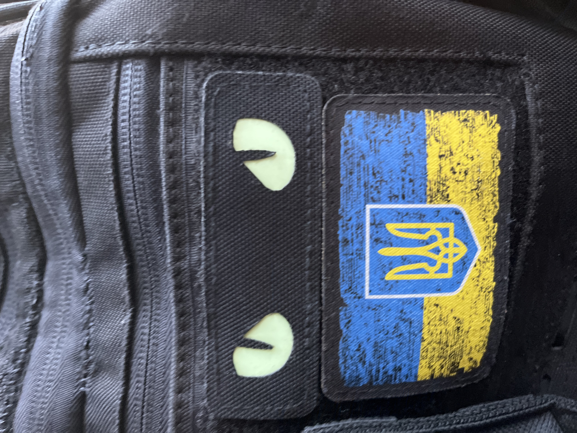 The image shows a close-up of a black tactical backpack with two patches attached. The top patch features two glowing, cat-like eyes. Below this, a second patch displays the flag of Ukraine with the national coat of arms in the center. The flag is rendered with a distressed, worn texture. The backpack itself has multiple zippered pockets and appears to be made of durable, textured fabric. The background is also composed of the same black, textured material of the backpack. The scene is devoid of any environmental context suggesting a specific location within Kriukivshchyna, Ukraine, or any other city. There are no people, actions, or interactions depicted.