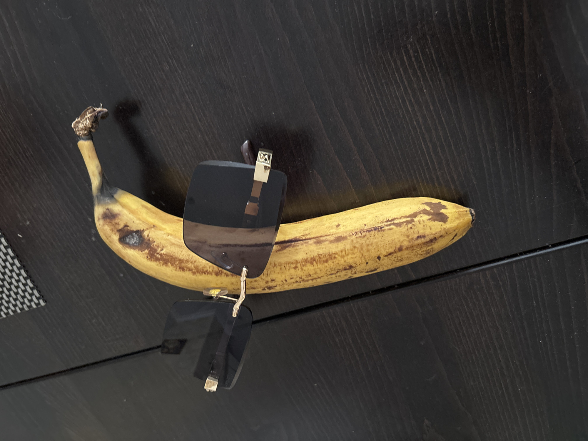 A slightly overripe banana with brown spots lies on a dark brown wooden surface. A pair of dark-lensed sunglasses is perched atop the banana. The sunglasses have silver-colored hinges. The scene appears to be indoors, possibly on a table. There is no indication of this scene being located in Budva, Montenegro.