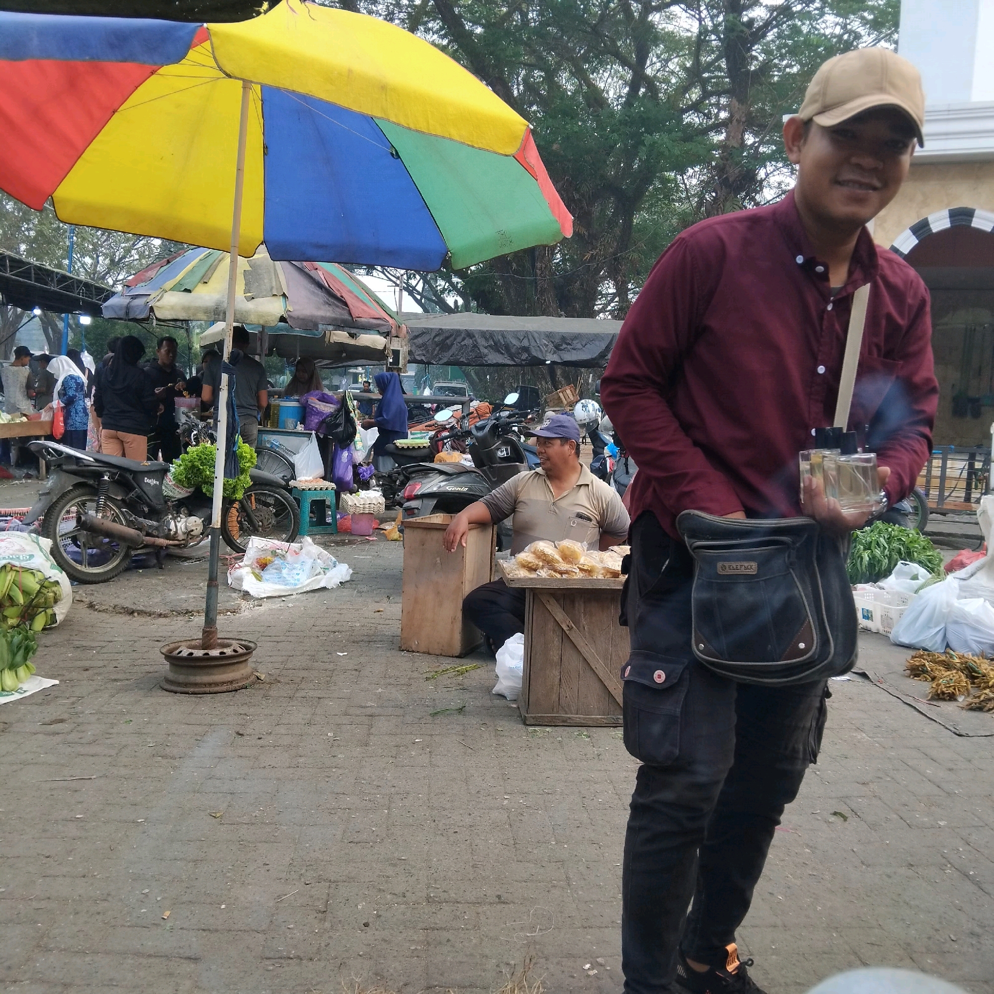 The image depicts an outdoor market scene in Banjarbaru, Indonesia. In the foreground, a man wearing a maroon collared shirt, black pants, and a tan baseball cap smiles at the camera while holding up two clear perfume bottles. He has a dark bag slung across his body. The background shows various market stalls with colorful umbrellas, including a large multi-colored umbrella partially covering the scene. Several people are visible at the stalls, some interacting and others engaged in their activities. A motorcycle is parked near a stall, laden with green leafy vegetables. The ground is paved with cobblestones, and there are scattered items such as bags of produce, crates, and trash. The overall environment suggests a bustling local marketplace with goods and people.