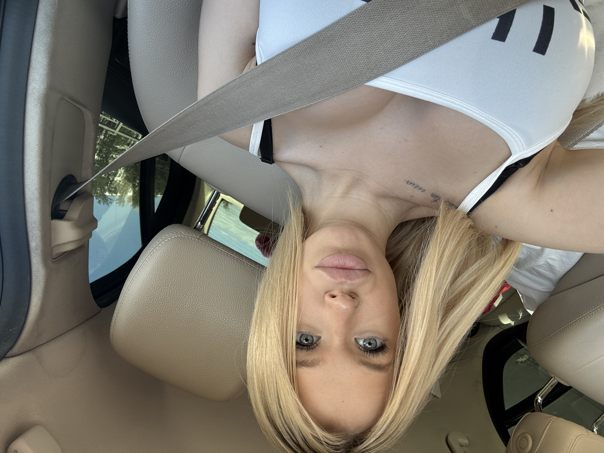 The media file is a portrait-oriented photograph taken inside a car. The main subject is a young Caucasian woman with blonde hair, blue eyes, and a nose ring. She is wearing a white tank top and a tan seatbelt crosses over her chest. The interior of the car appears to be made of tan leather and fabric. Sunlight is visible through the car windows, suggesting it is daytime. The woman's expression is neutral, and her eyes are looking directly at the camera. She has a tattoo on her collarbone. A portion of the car's headrest is visible in the foreground. The image quality is sharp, and the lighting is natural. The overall setting is consistent with a person taking a selfie while in a vehicle. There is no visible text in the image. The photo appears to have been taken in Saint Petersburg, Russia.