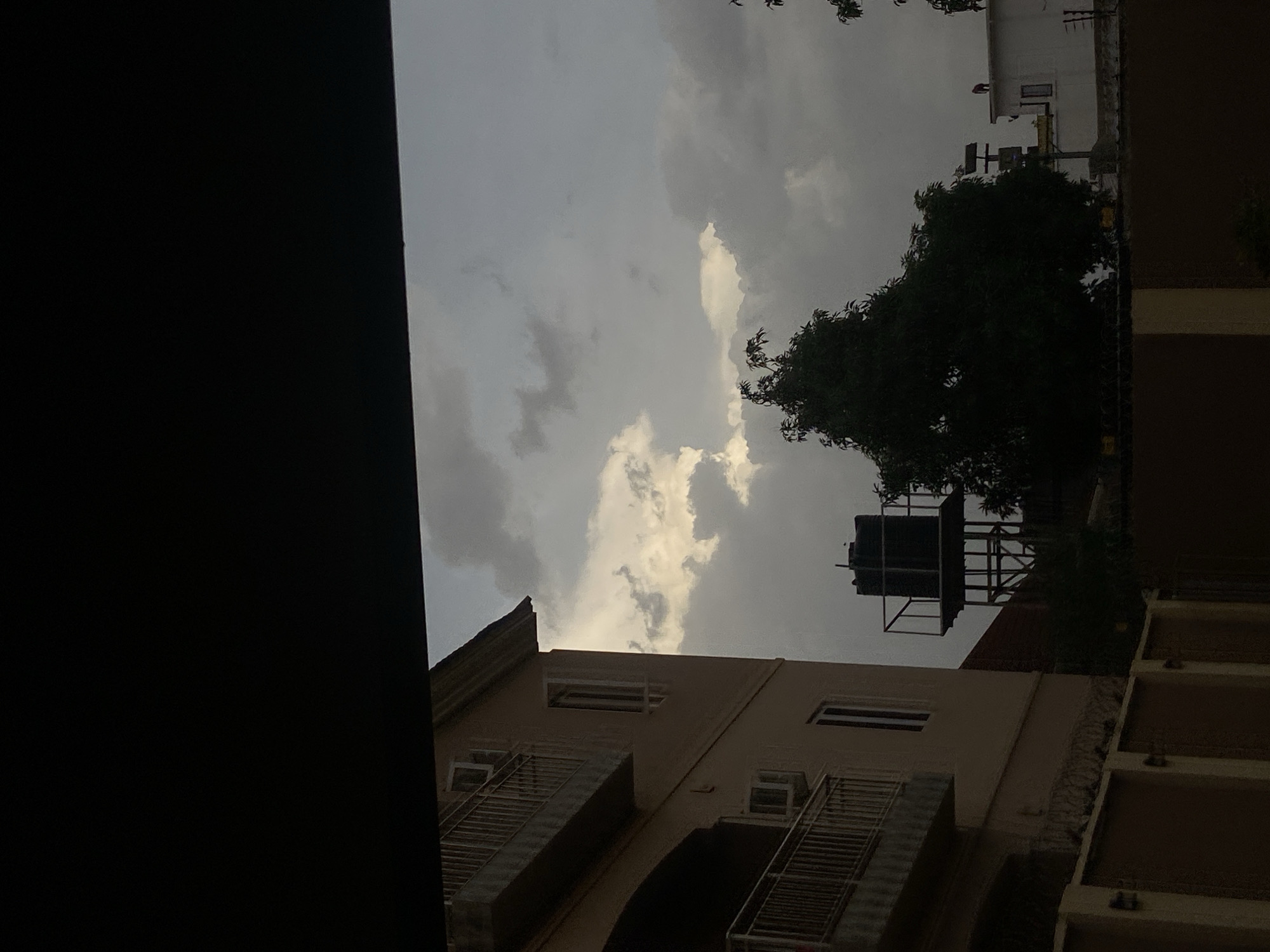 The image shows a view from inside a building in Barden Yana, Nigeria, looking out toward a cloudy sky. A portion of the building's window frame is visible on the left. The view includes buildings with balconies, a water tank structure, and a tree.