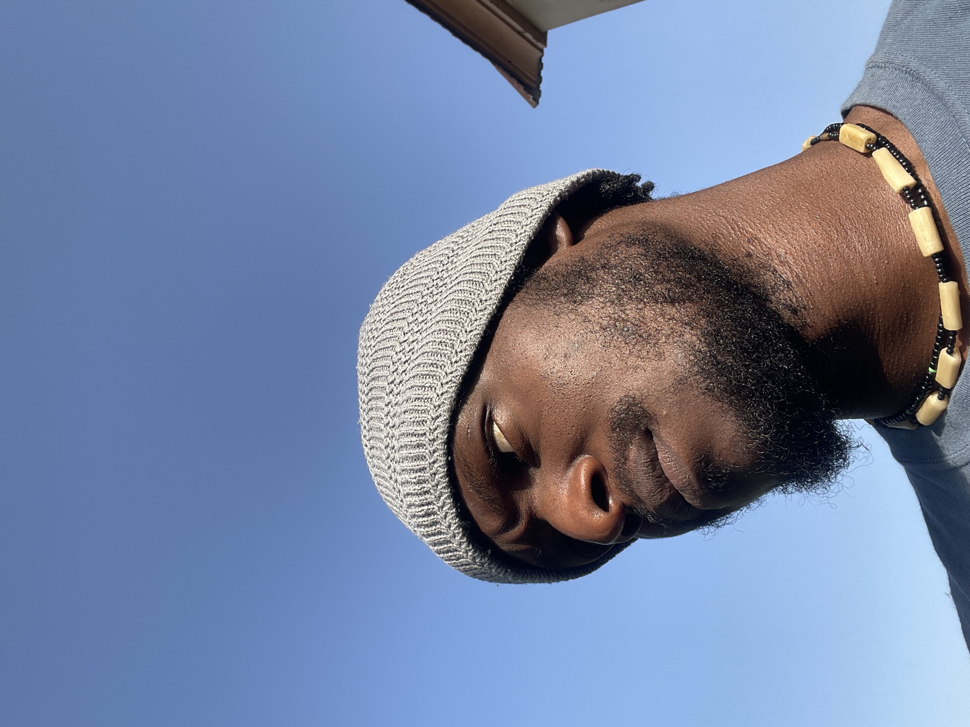 A low-angle shot of a Black male wearing a gray knit cap and a beaded necklace. The background is a clear blue sky. A portion of a building overhang is visible in the upper part of the frame. The location is likely Jos, Nigeria.