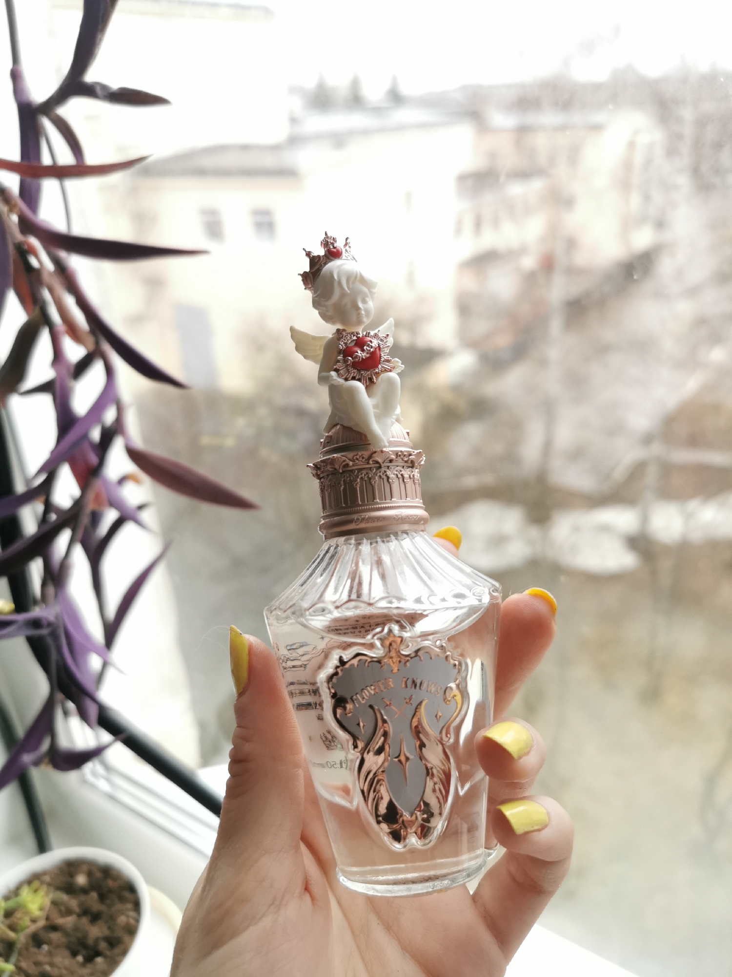 A hand with yellow painted nails is holding a clear glass perfume bottle with a small white angel figure on top. The angel figure wears a red heart and crown. The perfume bottle has a silver-colored label with text. A purple plant is partially visible in the left foreground and there is a plant in a white pot at the bottom left corner. The background is a blurry view of buildings seen through a window with what appears to be rain on the glass. Location: Unknown, Unknown.