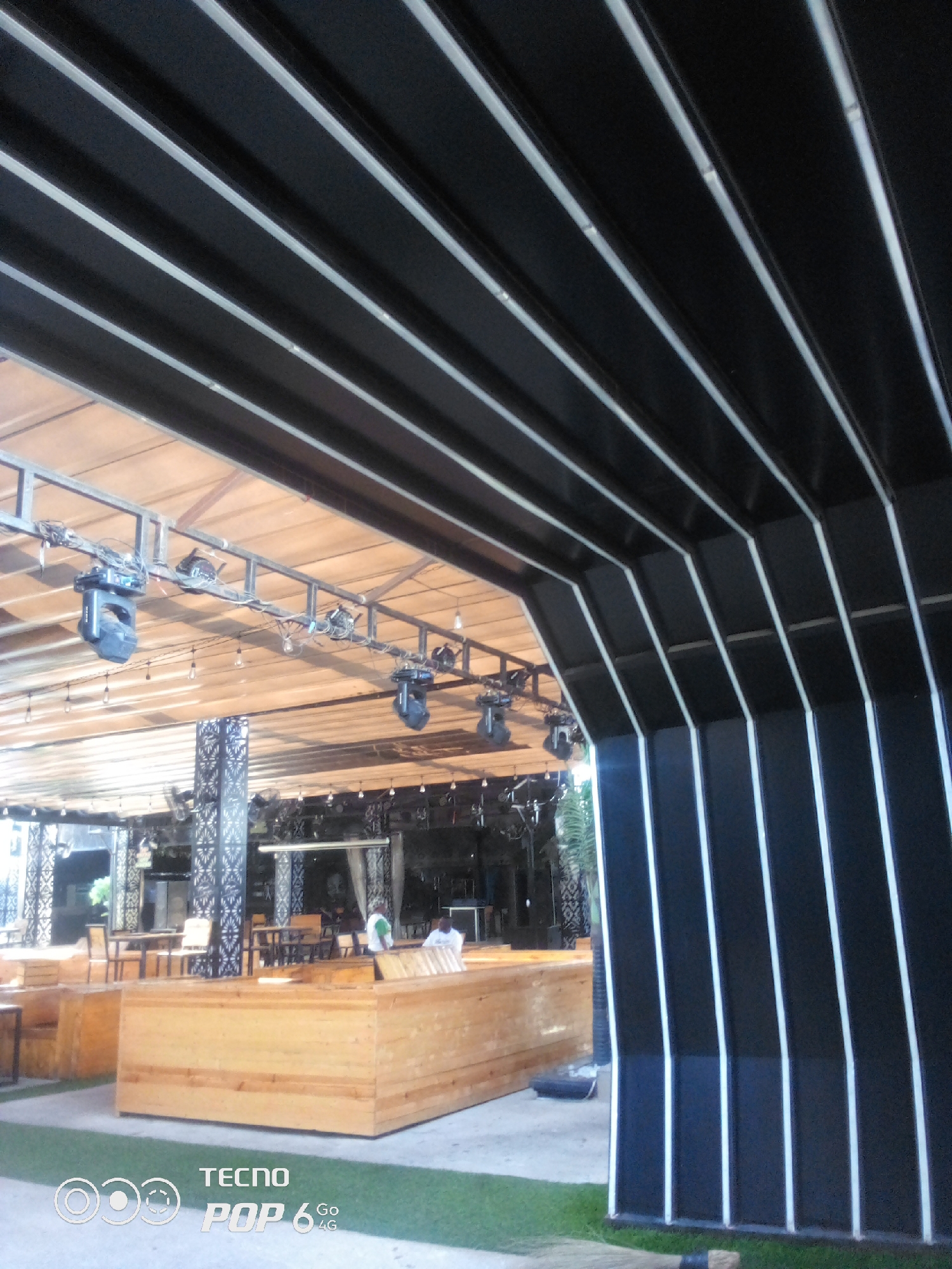 This media file depicts an outdoor seating area, likely a restaurant or bar, with a modern, black and white striped awning overhead. The awning consists of parallel black panels with thin white stripes, creating a sleek, contemporary design. Beneath the awning, a wooden ceiling is visible, supported by a metal frame from which several stage lights are suspended.
In the background, tables and chairs are arranged for patrons, suggesting a dining or social setting. Some of these tables appear to be made of dark wood, while others have lighter wooden chairs. The scene is furnished with wooden benches and low tables in the foreground, providing additional seating options. The ground is a mixture of concrete and grass.
There are a few people visible in the background, appearing to be staff or patrons. One person in a white shirt is seen standing near some tables, and another individual in a green shirt is also present. Their presence suggests that the venue is open and operational.
The lighting indicates it is daytime, possibly late afternoon or early evening, given the artificial lights being visible, but not necessarily in use. The weather appears to be fair, as it is an outdoor setting. The location is identified as Howayi, Nigeria. The lower part of the image contains a watermark with "TECNO POP 6 4G" printed on it.