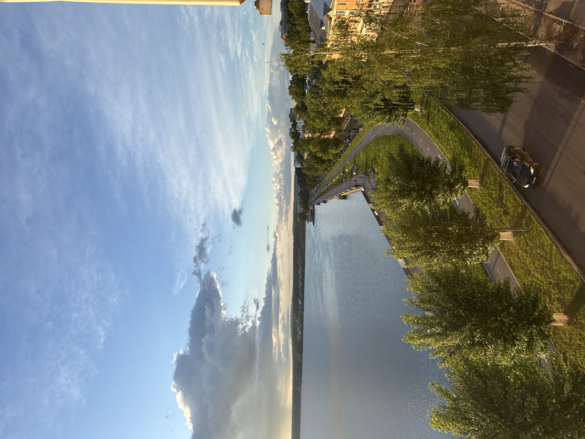 The image presents a riverside scene in Votkinsk, Russia. On the right, a road runs parallel to the river. A black car is on the road. A pathway separates the road from the waterfront, lined with trees. The river occupies the middle ground, reflecting the sky. The sky is partly cloudy with a mix of blue and gray clouds. Buildings can be seen along the opposite bank of the river.