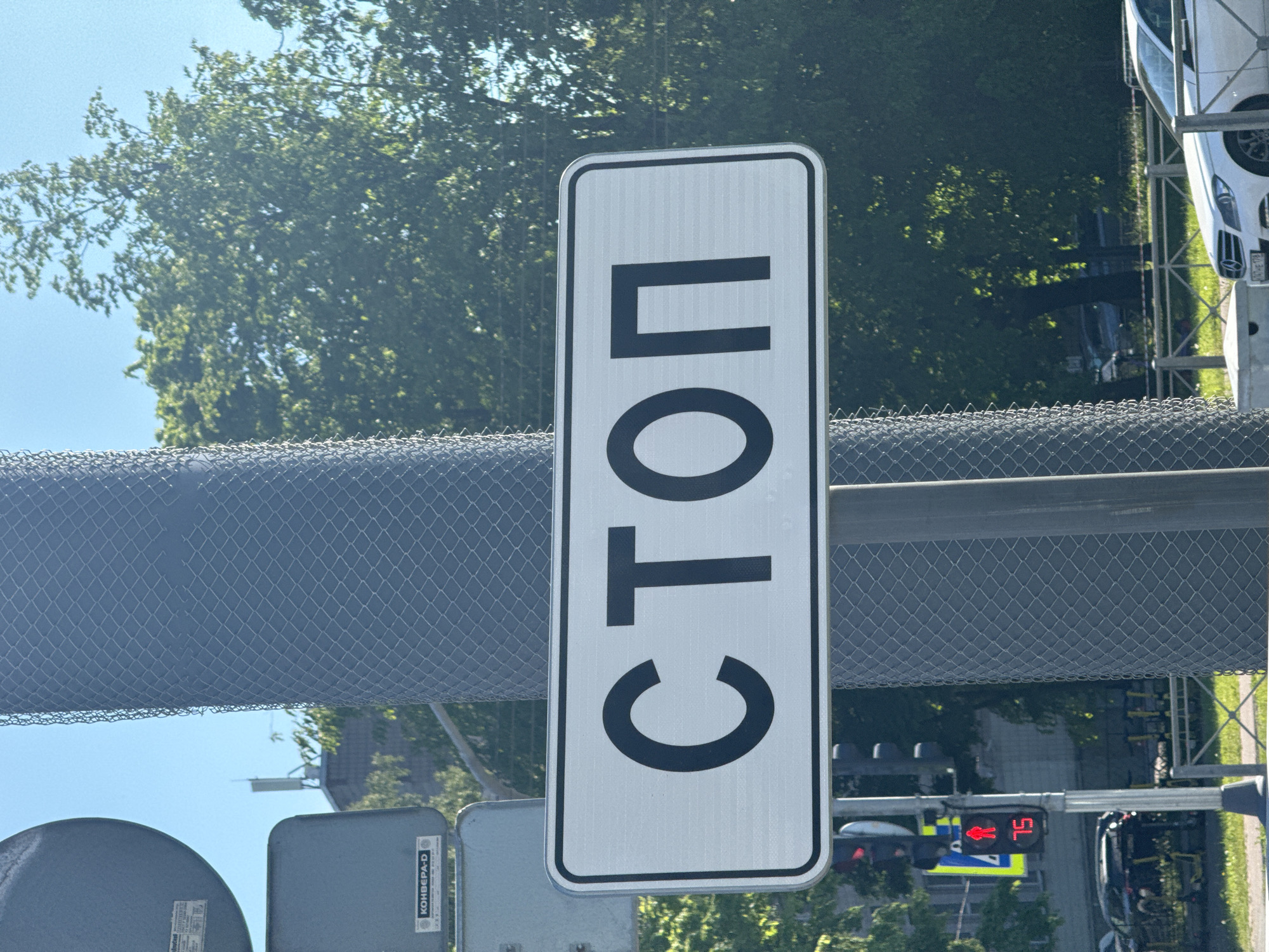 A brightly lit outdoor scene, captured during the day under a clear blue sky, displays a street or industrial area. The dominant feature is a tall, rectangular white sign with rounded corners, prominently featuring the Cyrillic word "СТОП" (STOP) in bold black letters, oriented vertically. The sign has a noticeable ribbed texture.
A grey chain-link fence stretches across the midground, acting as a barrier. Below the sign and fence, on the left, are several grey utility boxes. One box clearly shows the Cyrillic text "КОНВЕРА-D" and possibly "Redford" or "Radford".
In the background, abundant green trees suggest a treelined area or park. To the right of the main sign, a traffic light is visible, displaying a red pedestrian symbol along with what appears to be "5L" or "SL", possibly a countdown. A white car, likely an SUV, is partially visible behind the fence on the right side of the image. Faint structures or buildings can be seen in the distance through the foliage.
No specific activity or people are shown; the image focuses on urban infrastructure. The Cyrillic text strongly indicates a location in Russia. The bright daylight suggests a sunny day.