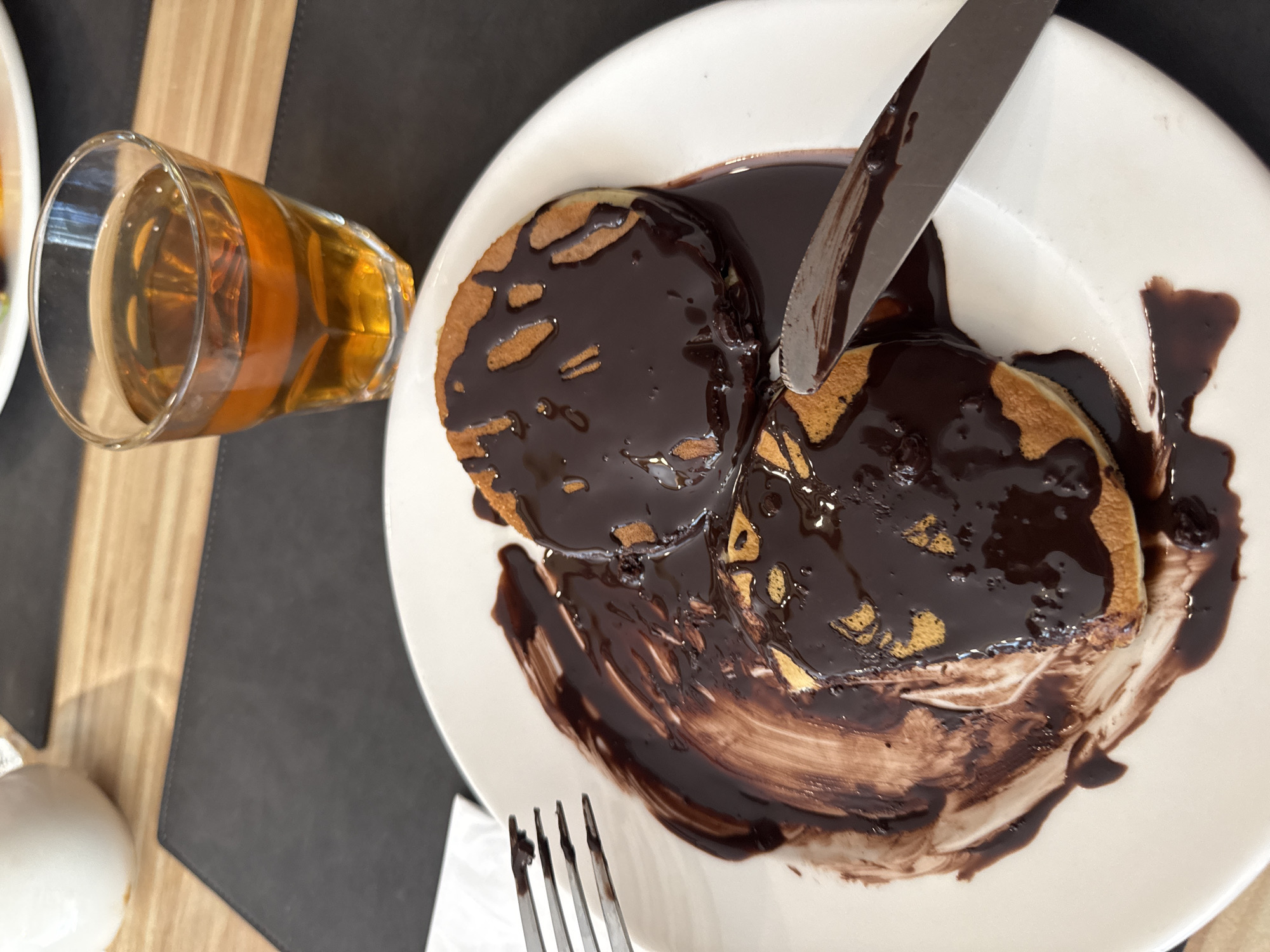 The image shows a plate of chocolate-covered pancakes, a glass of amber liquid, and cutlery on a table. The pancakes are small and round, with a dark chocolate sauce generously poured over them. The chocolate sauce is also spread around the plate, creating a messy yet appealing presentation. A fork is positioned to the left of the plate, with some chocolate sauce on its tines. A knife rests on the right side of the plate, also with some chocolate sauce on it. The glass contains a beverage that appears to be tea or a similar amber-colored liquid. The table surface is a light wood grain, covered in part by a dark placemat. A white pitcher or creamer is partially visible in the upper left corner of the image. The overall scene suggests a breakfast or dessert setting in a restaurant or cafe. There are no people visible in the image. The scene is an indoor shot, likely in Aksu, Turkey, given the prompt.