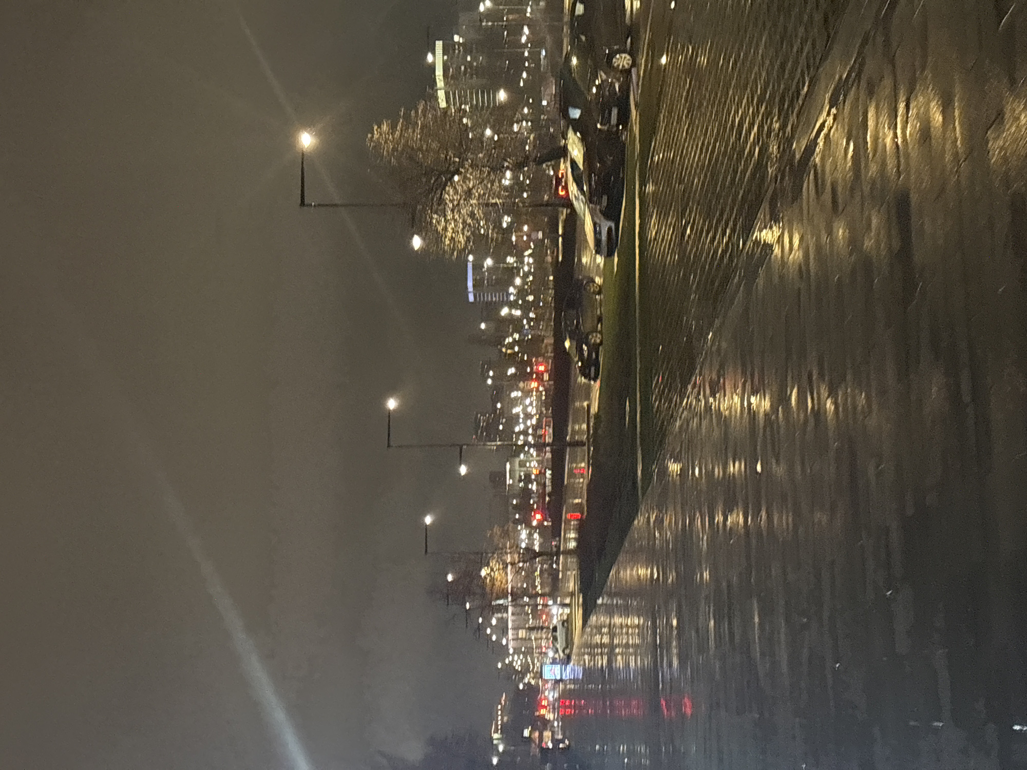The image depicts a rainy night scene on a street in Perm, Russia. Streetlights illuminate the wet pavement, reflecting the city lights. Parked cars are visible along the curb. The atmosphere suggests a cold, damp evening. The image captures a typical urban night scene.