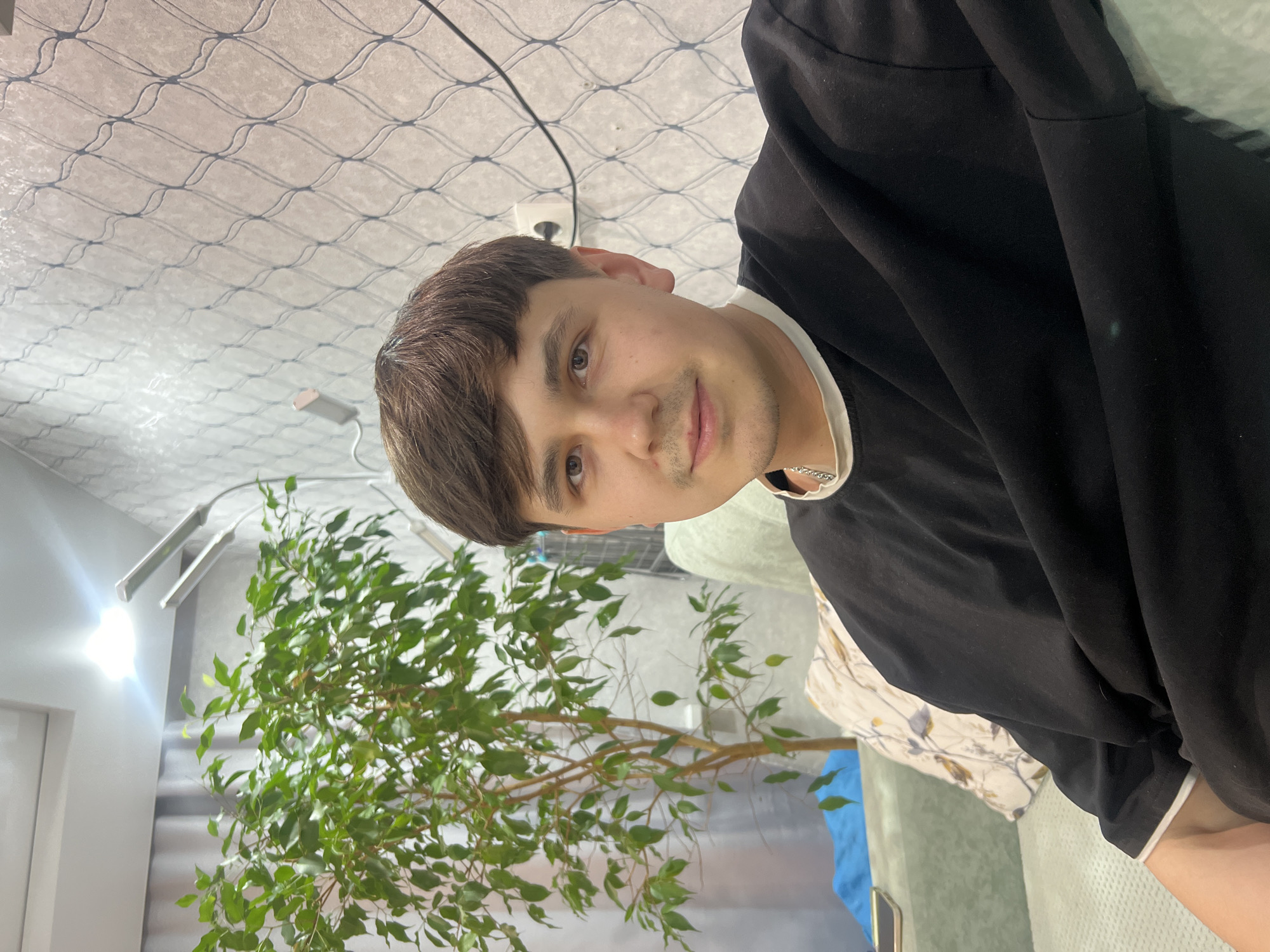 A person wearing a black long-sleeve shirt and a white undershirt is in an indoor setting, possibly a room in a home or office. The person has brown hair and light skin. In the background, there is a faux tree, blue drapery, a ceiling with a patterned texture, and a wall-mounted light fixture. The location is potentially in the city of Chaikovsky, Russia.