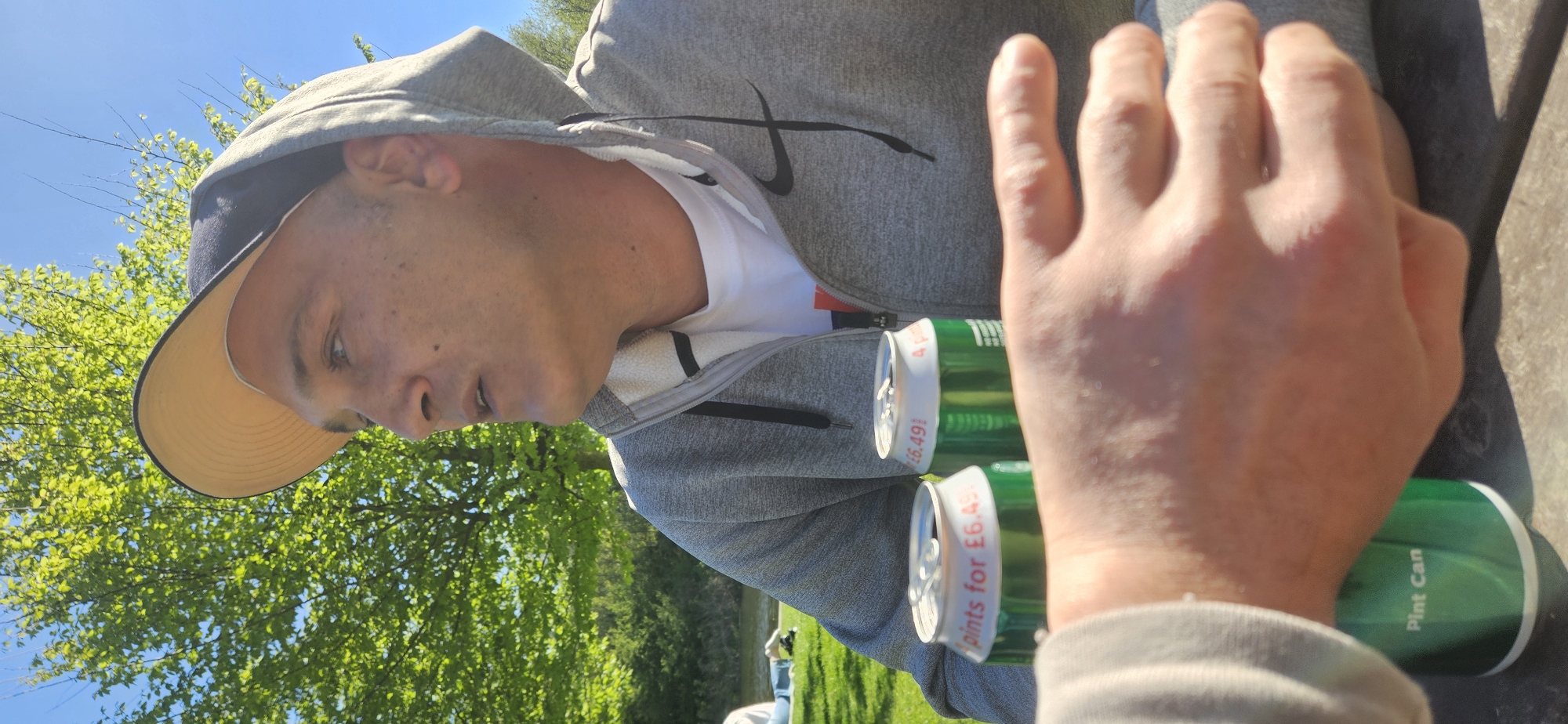 The image captures a casual outdoor scene on a sunny day, likely in a park or open green space. The primary subject is a male, seen from a slightly elevated, side-angle perspective. He wears a grey hooded sweatshirt with a visible black Nike swoosh logo on the chest, a white t-shirt underneath, and a dark cap with a tan-colored brim. His head is turned slightly to his right, and his gaze appears directed downwards or towards something out of frame.
In front of him, two green cans with white tops are visible. Text on the cans clearly reads "Pints for £6.49" and "Pint Can," indicating they are alcoholic beverages and providing a price point. A person's hand, presumably belonging to another individual or the photographer, is prominently featured in the foreground, close to the cans. This hand is illuminated by bright sunlight, casting slight shadows.
The background features lush green trees with bright foliage against a clear, vibrant blue sky, suggesting a warm and pleasant day. Further in the background, a grassy area is visible, with another person faintly seen in the distance, indicating a social gathering or public park setting. The strong sunlight creates clear shadows and highlights the textures of the trees and clothing. The visible text on the cans, particularly the currency (£), aligns with the provided context of Norwich, United Kingdom.
