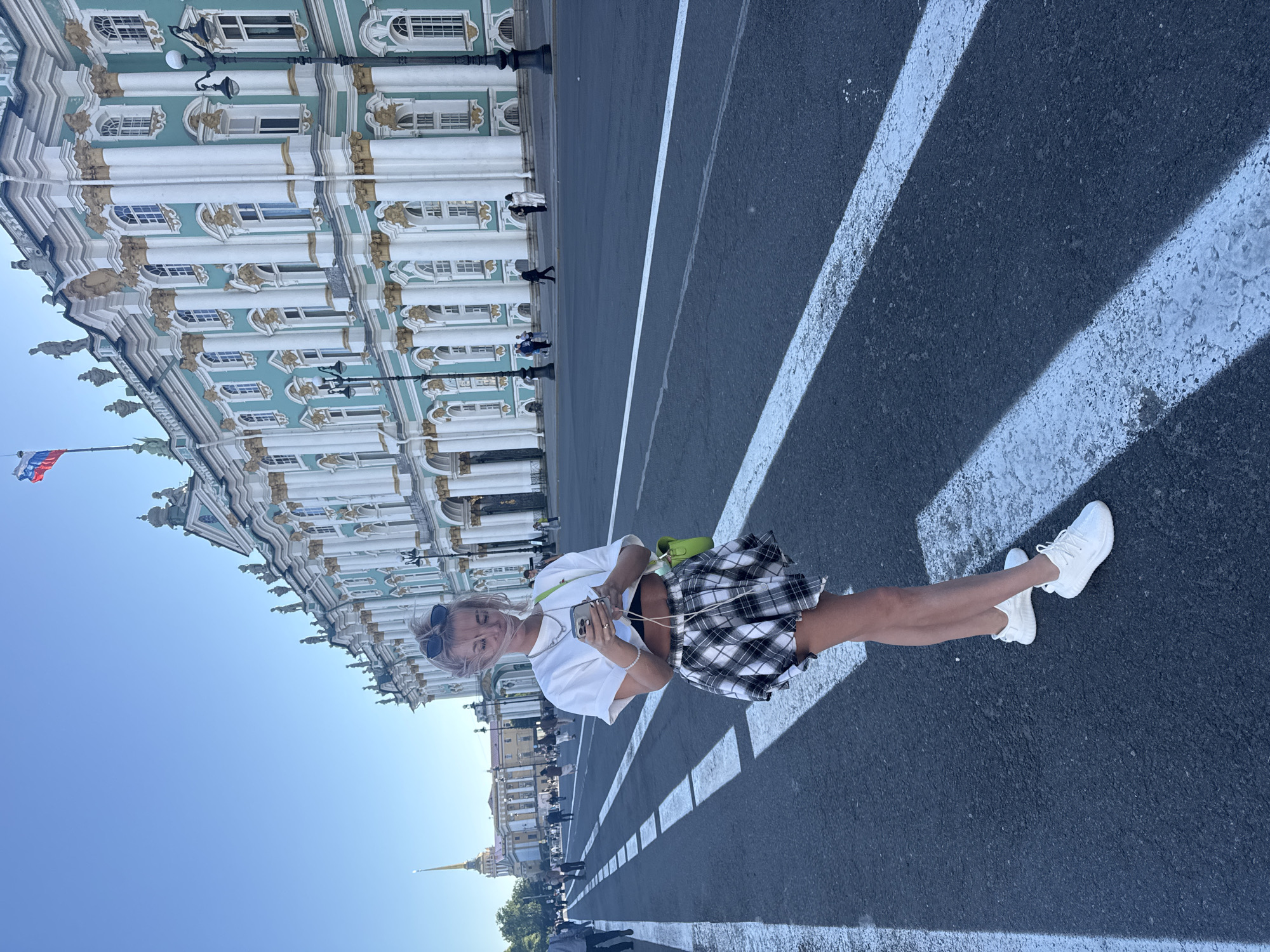 A young woman with blonde hair is standing on a street in Saint Petersburg, Russia, looking at her phone. She is wearing a white crop top, a black and white plaid skirt, and white sneakers. She has a bright green bag slung over her shoulder and sunglasses perched on her head. The iconic Winter Palace, painted in a striking turquoise and white, serves as a grand backdrop, its elaborate architecture under a clear blue sky. The street in front of the palace is wide, with white pedestrian crossing lines painted on the asphalt. In the distance, the spire of the Admiralty Building can be seen. Other pedestrians are scattered across the square, some walking, others standing. The overall scene depicts a sunny day in a historic and bustling urban environment.
