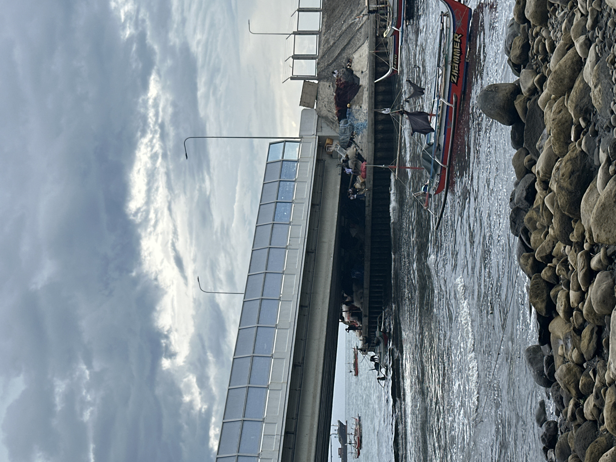 The image depicts a cloudy day in Davao City, Philippines, near a bridge or overpass. In the foreground, a rocky shore leads to choppy, dark gray water. Several small, colorful fishing boats are scattered on the water, some bobbing close to shore and others further out. One boat in the middle ground features red and white pontoons and has a black flag flying from its mast. The name "ZHÄMMER" is visible in red lettering on the side of this boat. In the mid-ground, under the overpass, are several people, piles of fishing nets, and what appear to be storage materials. Large ships can be seen in the distance on the horizon. The overpass itself is a modern structure with a white railing and tinted glass panels. The overall scene suggests a working harbor or coastal area.