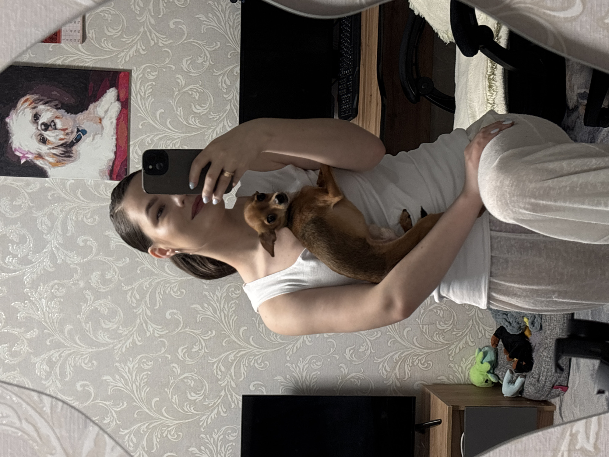 A woman is taking a selfie in a mirror with a small brown dog nestled against her chest. She is wearing a white tank top and light-colored pants. She holds a cell phone up to her face with her left hand and the dog is nestled between her left arm and her chest. A painting of a white dog with a pink bow is hung on the wall next to a calendar. The wallpaper is a light gray with a floral pattern. There is a television and a wooden cabinet in the background. There are stuffed animals sitting on the cabinet. A chair is visible behind the woman. The location is in Chaikovsky, Russia.