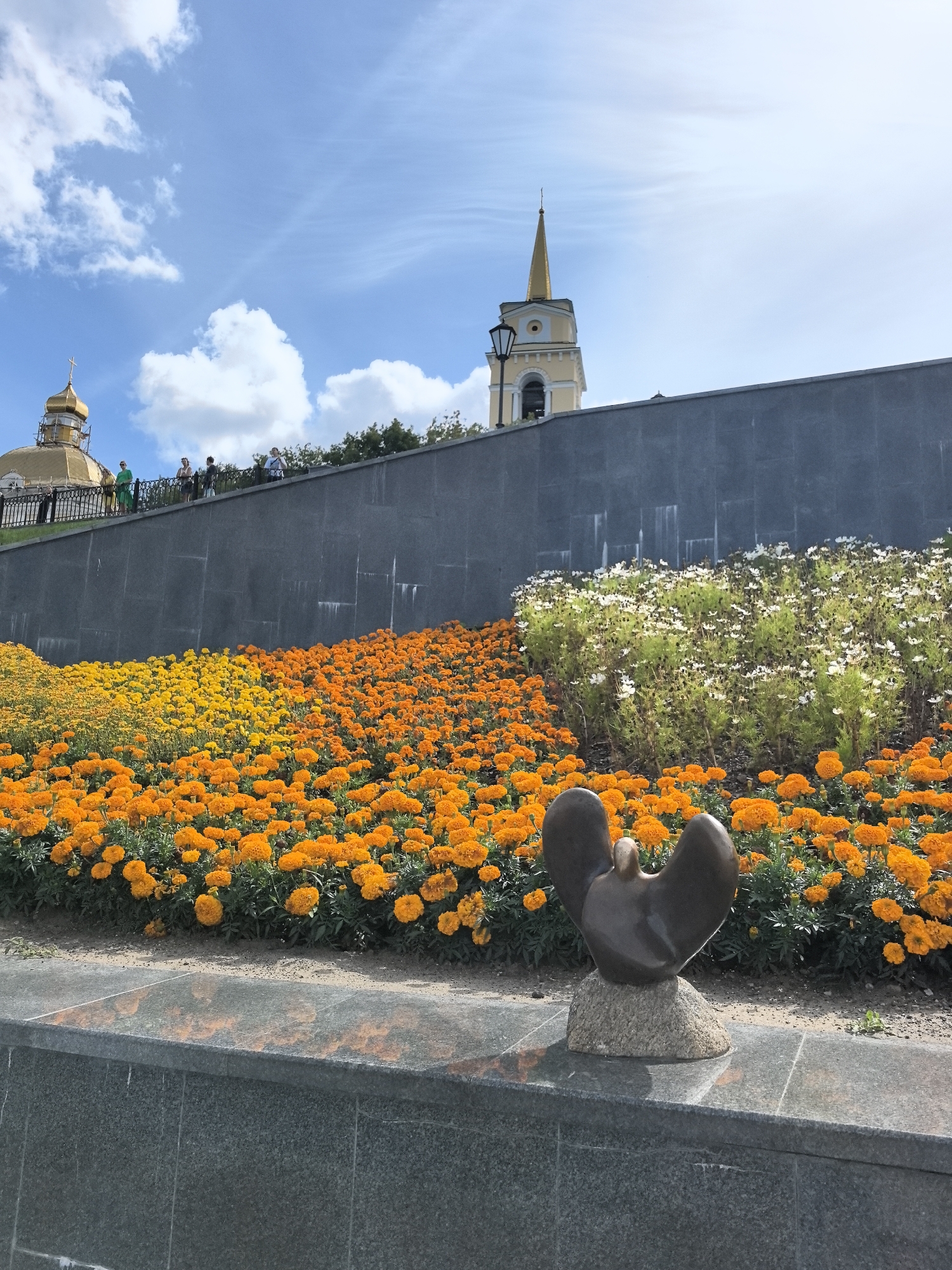 The image depicts a public space in the city of Perm, Russia, featuring a vibrant flower bed of orange and yellow marigolds, interspersed with white flowers. A small, dark bronze sculpture resembling a bird with outstretched wings rests on a stone base in the foreground. Behind the flowers, a dark gray stone wall rises, topped by a walkway where several individuals are visible. Beyond the wall, a golden-domed building with a cross, likely a church, and a tall, slender yellow bell tower with a gold spire are prominent architectural features against a blue sky dotted with white clouds. The scene suggests a sunny day in a well-maintained urban environment.