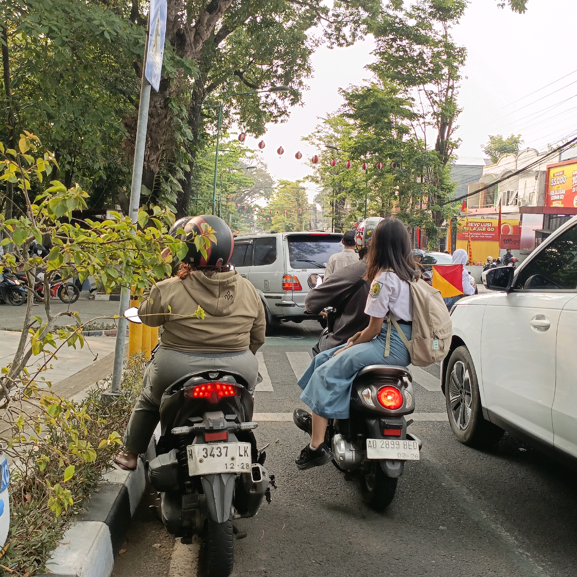This image captures a street scene in Salatiga, Indonesia, likely during daylight hours. The foreground is dominated by two motorbikes waiting in traffic. The motorbike on the left is ridden by a person wearing a hooded jacket and a helmet with a colorful design. Their legs are clad in grey pants. The motorbike on the right is carrying two individuals. The rider, wearing a light-colored shirt, is partially obscured. The passenger, a young person in a school uniform consisting of a white polo shirt and a blue skirt, has a beige backpack slung over their shoulder.
The motorbikes are positioned on a paved road, with white lines indicating a pedestrian crossing. To the left of the road is a curb with a low-lying green bush. A traffic signpost stands near the curb.
In the background, more traffic is visible, including a silver minivan and a white car. Buildings line the street, some featuring advertisements. Red and white decorative lanterns are strung across the road, suggesting a festive occasion or cultural event. Trees with lush green foliage are abundant, casting shadows and creating a canopy over parts of the street. The sky is overcast, with a hazy quality to the light. The overall impression is of a busy urban environment.