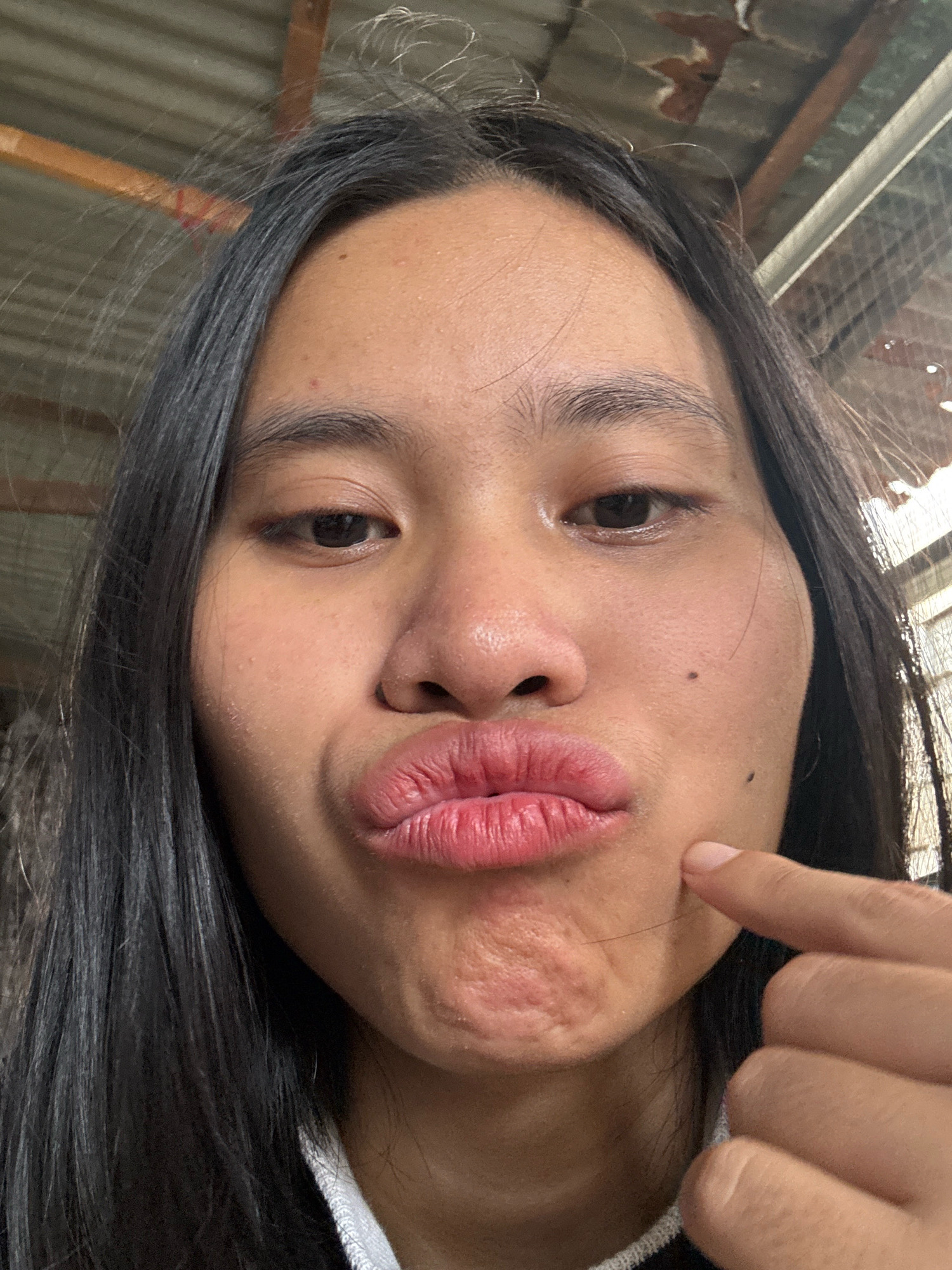 A close-up, waist-up selfie of a young Asian woman with long, dark hair, taken outdoors. She is making a kissing face, with her lips pursed. Her right index finger is touching her cheek. The background appears to be a corrugated metal roof and a chain-link fence, suggesting a simple, possibly rural or semi-urban setting. The lighting indicates daylight. The image was taken in East Jakarta, Indonesia.