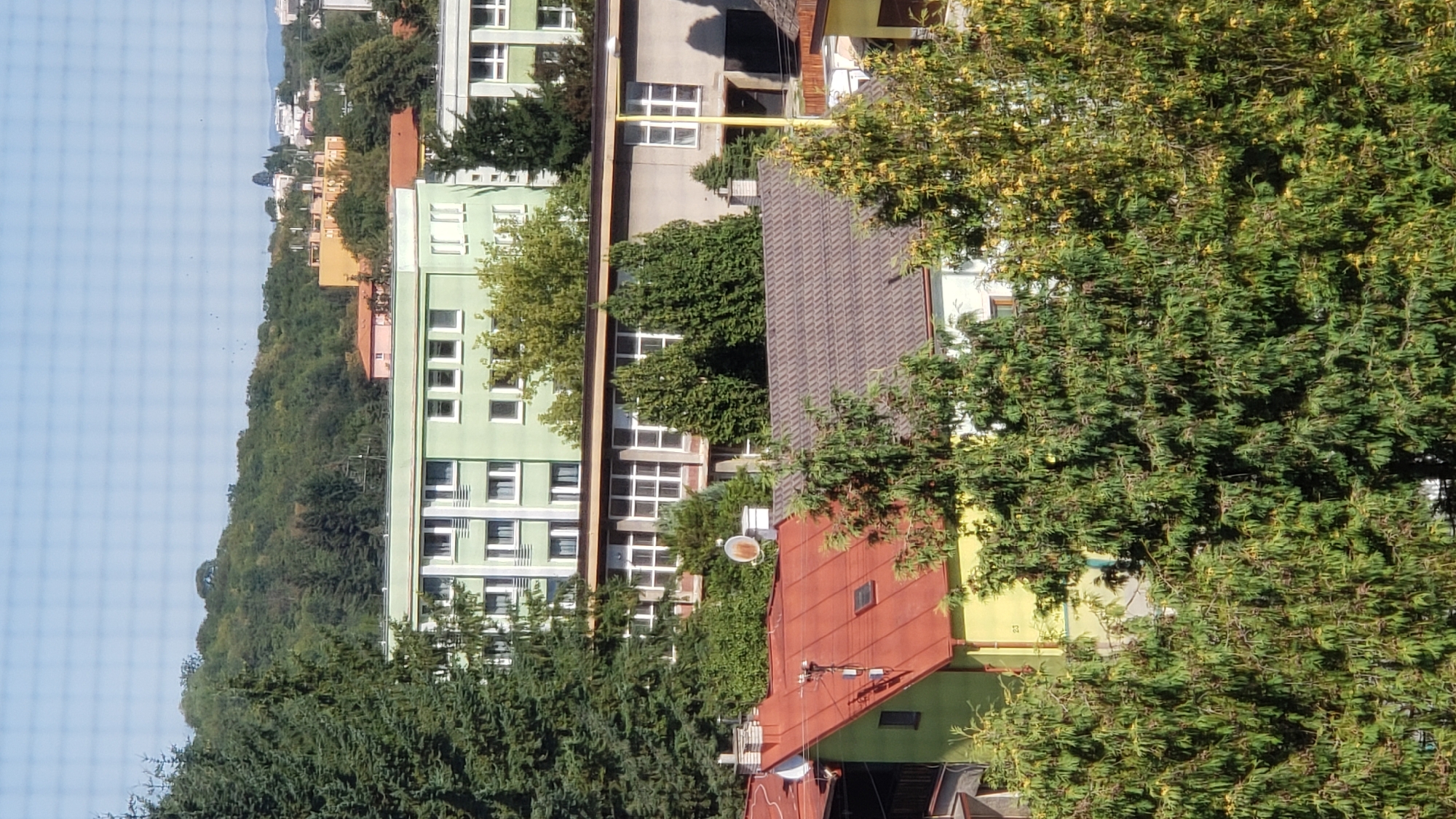 The image captures an aerial view of a residential area in the city of Prešov, Slovakia. In the foreground, dense green trees partially obscure the view of several buildings. A prominent red-roofed house with a yellow facade is visible on the left, with a smaller house featuring a dark, textured roof behind it.
Further back, a light green, multi-story building with numerous windows dominates the mid-ground. To its right, a longer, grey building with a facade of windows is partially visible. Beyond these buildings, a densely wooded hillside rises, with several scattered buildings and structures visible in the distance, including what appears to be a yellow and pink structure. The sky is a clear, light blue. The scene is composed of residential buildings and natural foliage, indicative of an urban or suburban environment.