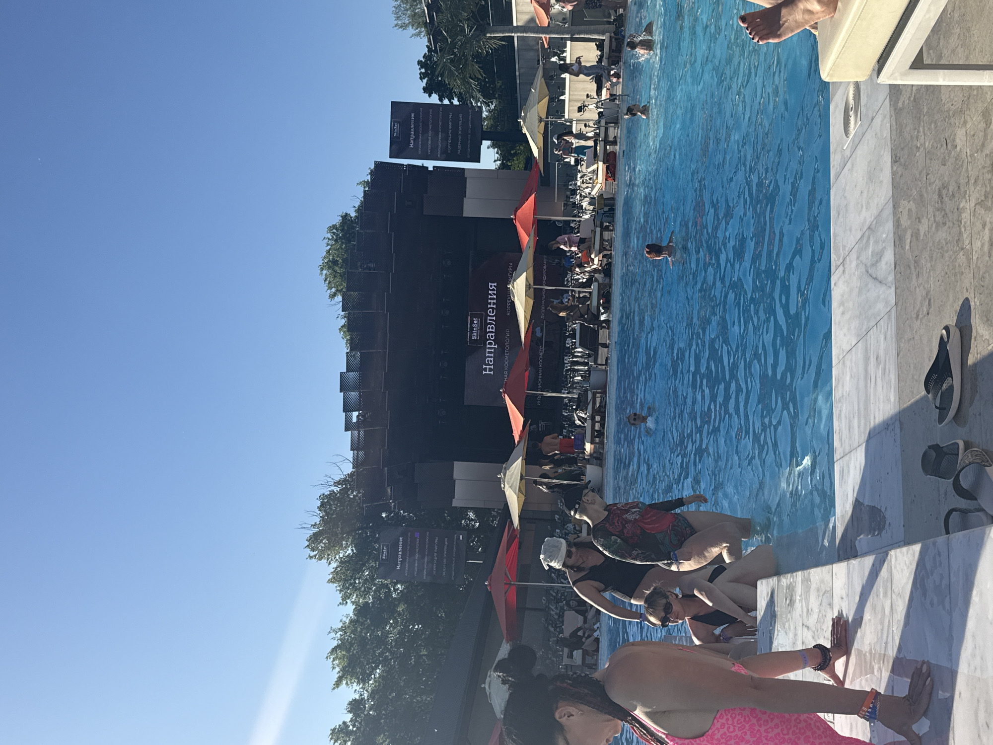 The image shows a swimming pool scene in Rostov-on-Don, Russia. The pool is surrounded by people swimming and relaxing. A large sign with Russian text, likely advertising something related to cosmetology, is visible in the background. There are lounge chairs and umbrellas near the pool. The sky is clear and blue. People are entering the pool near the edge of the pool.