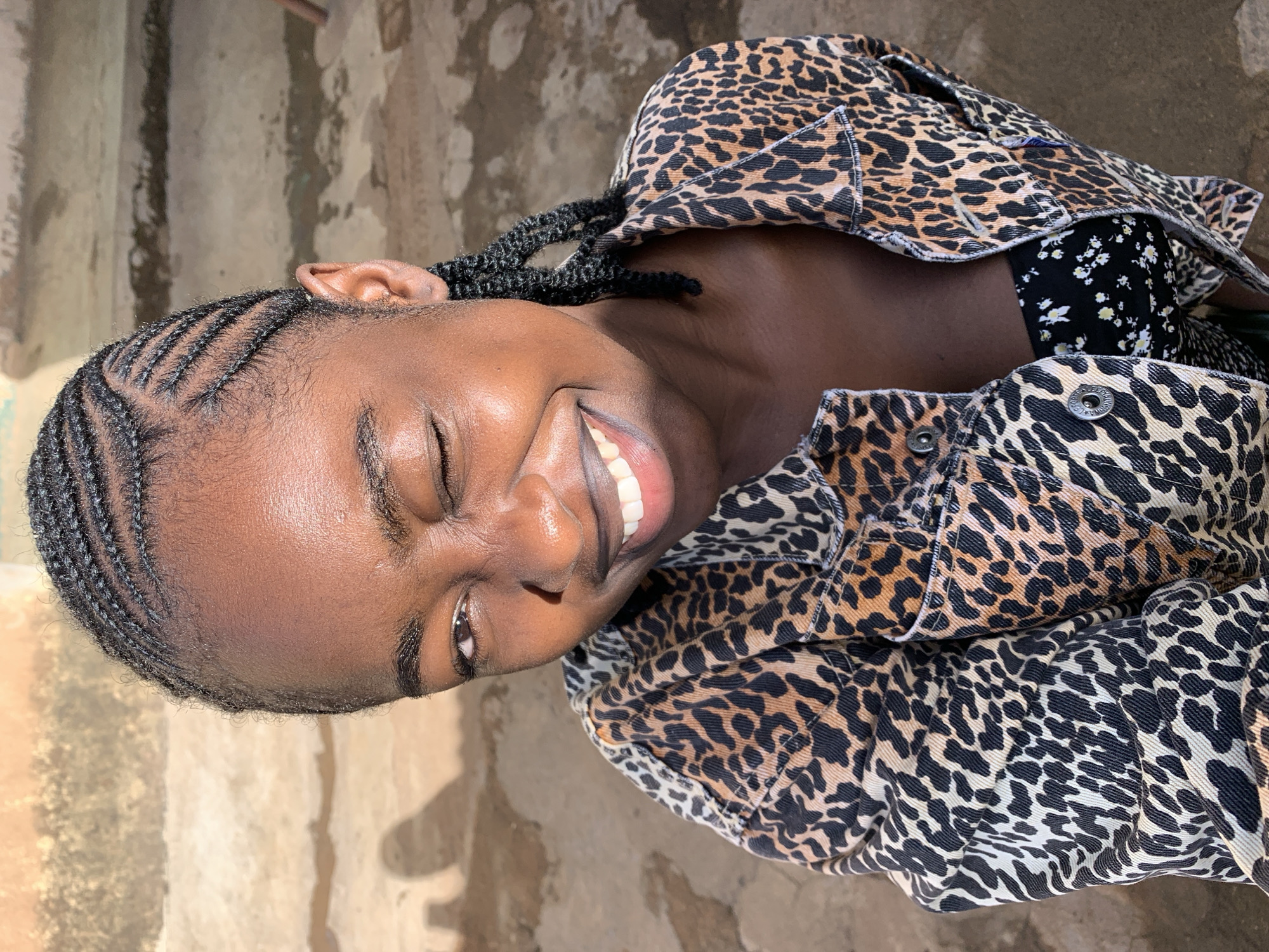 A close-up portrait captures a young person, likely female, with dark skin, smiling joyfully outdoors in Rafin Par, Nigeria, during daylight hours. Her head is tilted, and she gazes directly at the camera with a wide, radiant smile, her eyes squinted in happiness and white teeth visible. Her hair is styled in intricate cornrow braids that trace neat patterns across her scalp. She is wearing a prominent leopard print denim jacket, which appears to be layered over a dark shirt adorned with small white floral patterns. Natural light illuminates her face, giving her skin a subtle sheen. The background features a textured, light-colored wall or structure and a concrete or dirt ground, indicative of an outdoor setting. No clearly legible text is visible on her attire or in the immediate surroundings.