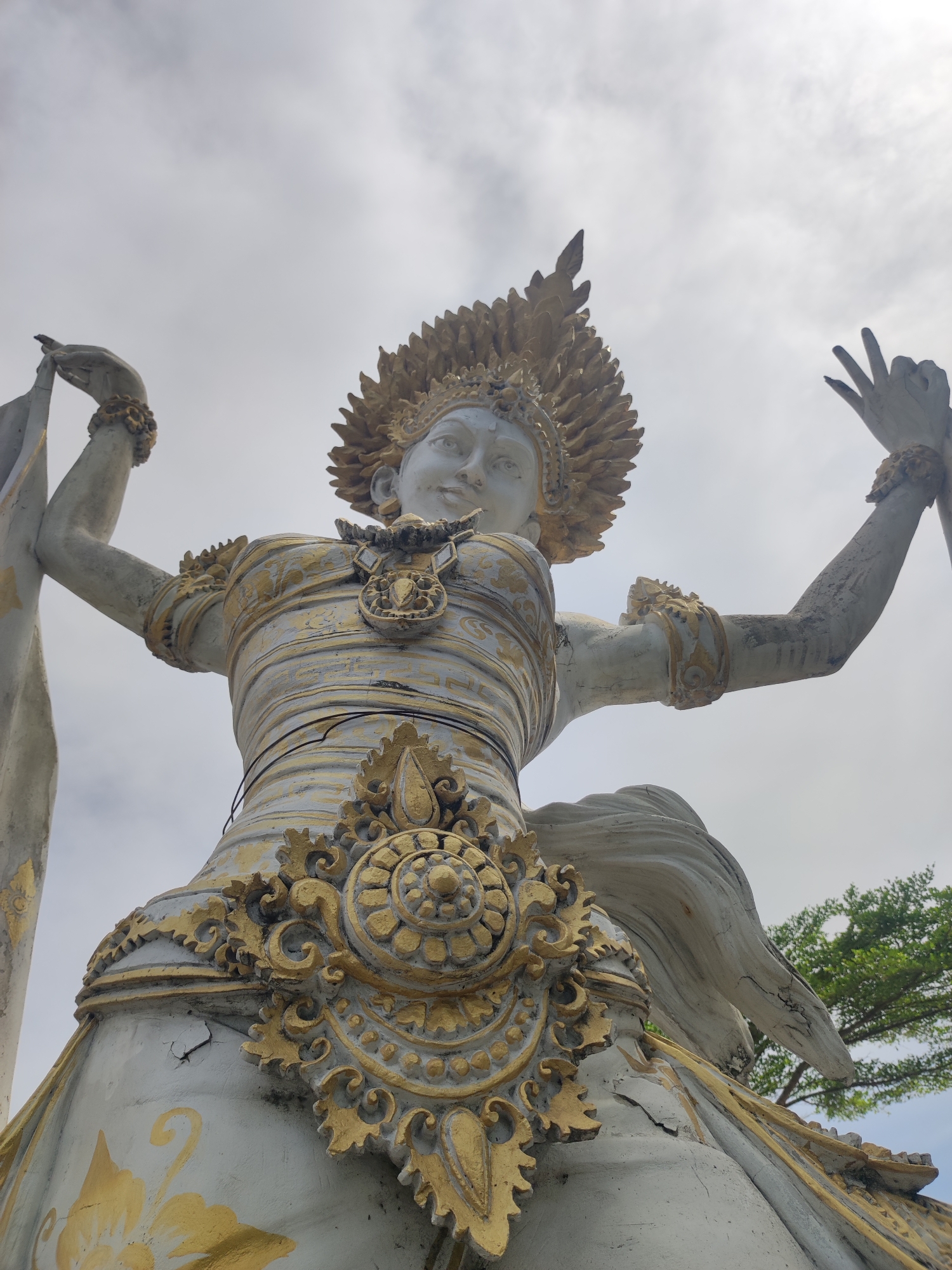A low-angle, full shot depicts a Hindu deity statue against a backdrop of a pale, overcast sky in Unknown, Indonesia. The statue, predominantly white with gold accents, features a female figure adorned with elaborate jewelry, including a large pendant and armbands. A spiky, golden headdress crowns her head. Her arms are raised, and her hands are posed in traditional mudras. A piece of white fabric or garment drapes from her left hand.
The statue's attire is ornate, with gold detailing highlighting the folds and design. Cracks and weathering are visible on the statue, indicating age and exposure to the elements. To the right of the frame, a glimpse of green foliage from a tree can be seen.