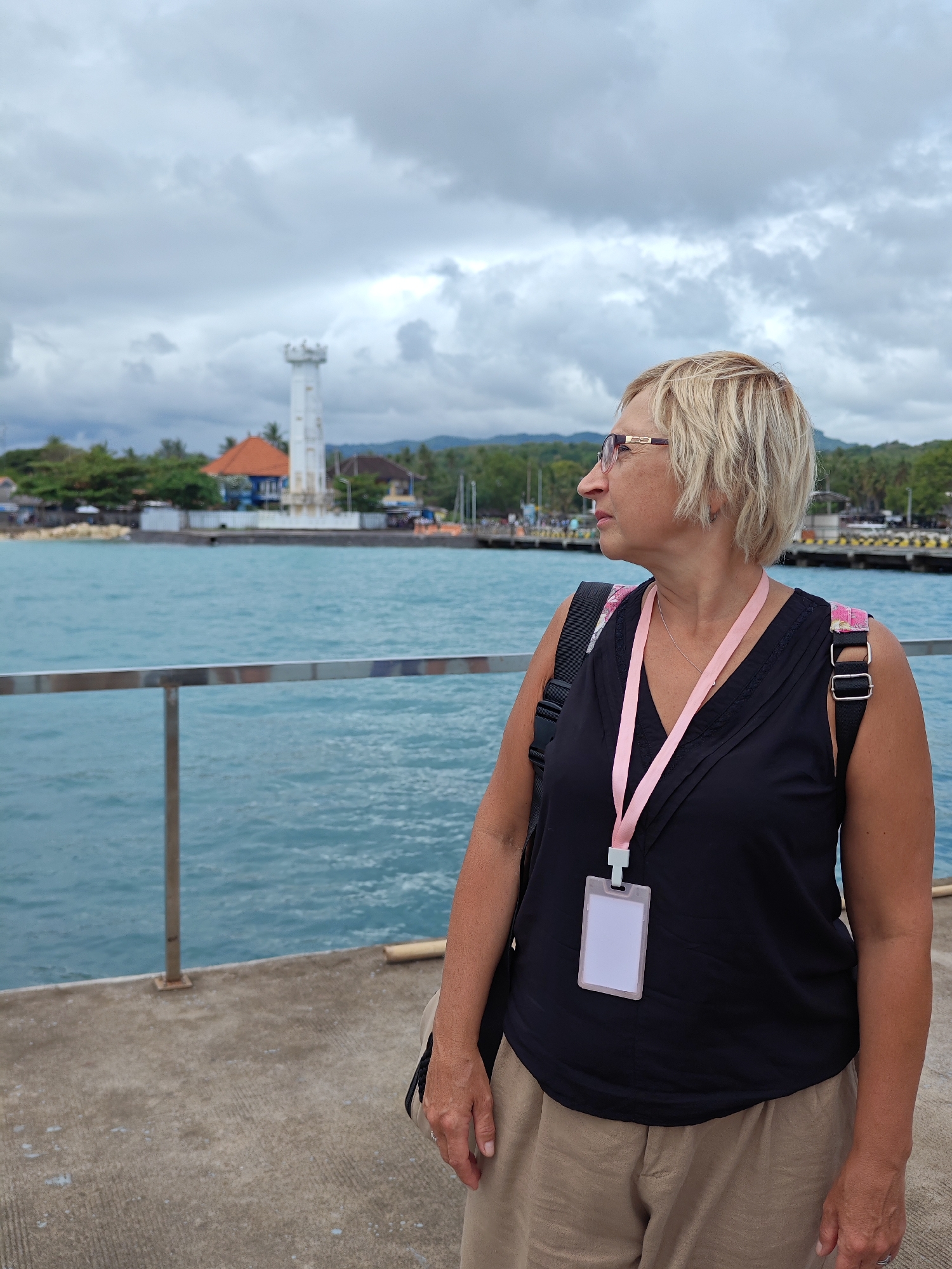 The image shows a middle-aged woman with blonde hair and glasses standing on a concrete pier in Kampung Toyapakeh, Indonesia. She wears a black sleeveless top, tan pants, and a pink lanyard with an identification badge. A black backpack hangs on her shoulder. She is looking to her right, towards the water. Behind her is a view of the ocean, with a white lighthouse on a small island. The sky is cloudy. A railing is visible along the pier.