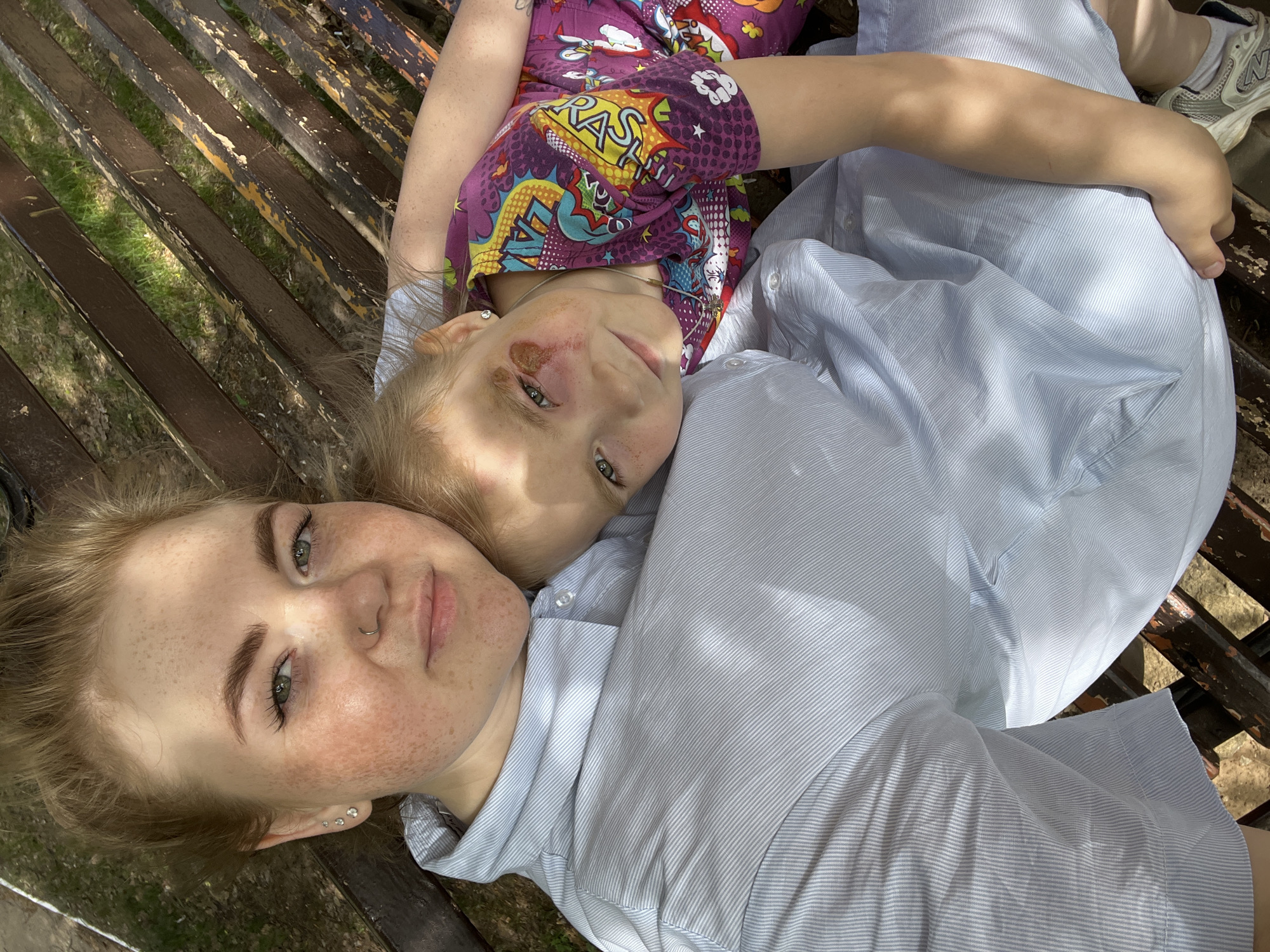 Two individuals, one adult and one child, are recumbent on a slatted wooden bench. The adult, positioned at the bottom of the frame, has fair skin with freckles, light hair, and wears a light blue, striped button-down shirt. The child lies atop the adult, wearing a purple t-shirt with cartoon-like graphics. A visible abrasion is present near the child's eye. The bench, composed of weathered wooden slats, is located outdoors, evidenced by glimpses of greenery visible between the slats. A shoe is visible in the upper right corner of the frame. The location is the city of Chaikovsky, Russia.