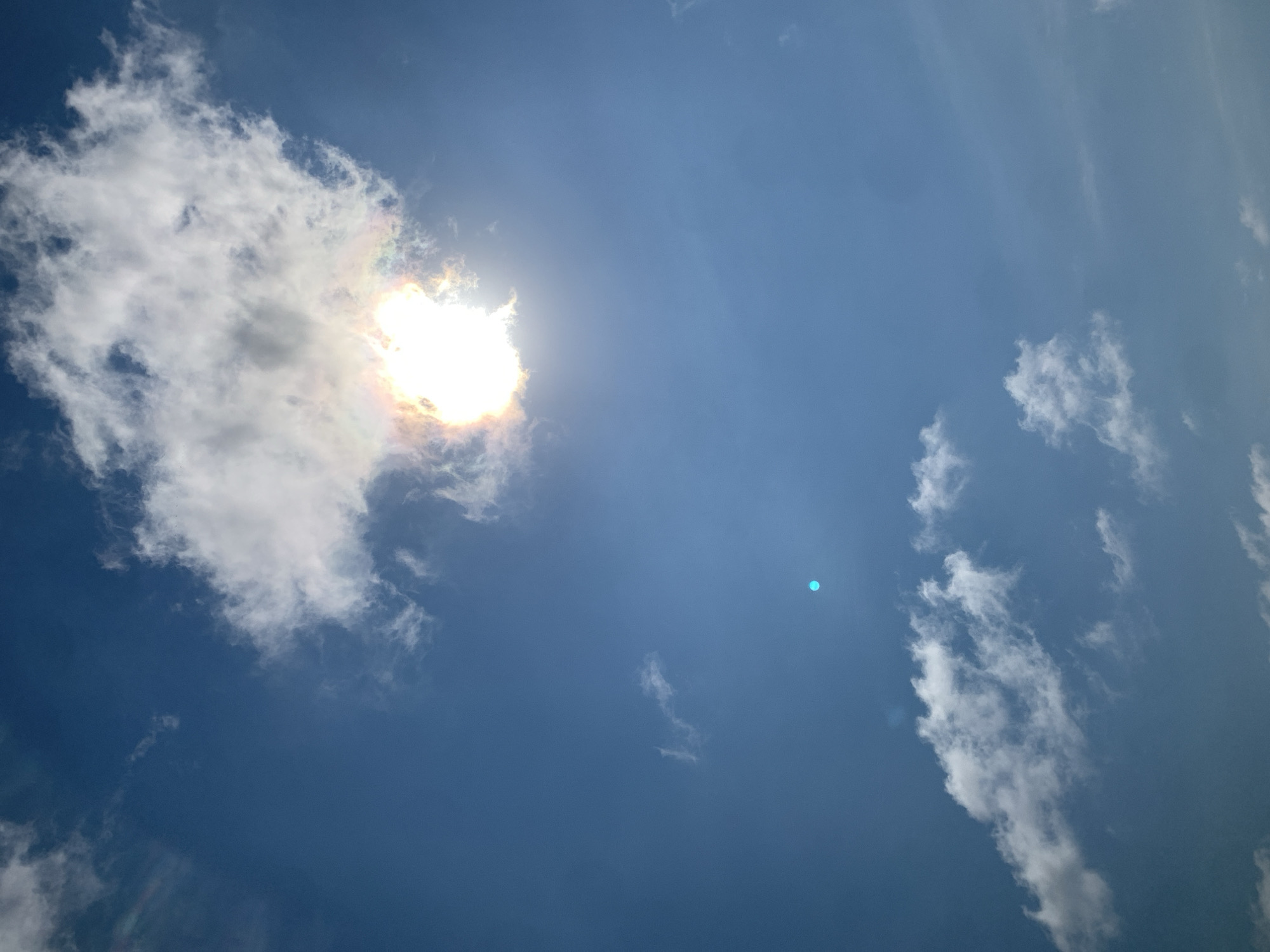 A clear blue sky dominates the image, with scattered white clouds. The sun is prominently displayed, partially obscured by a fluffy cloud, creating a bright, almost halo-like effect with iridescent colors. In the mid-ground, a small, distinct turquoise circle is visible. The lower portion of the image features more scattered clouds, some appearing wispy and others more dense. No people, buildings, or specific landmarks are visible, making it impossible to determine a precise location within Khazar Raion, Azerbaijan.