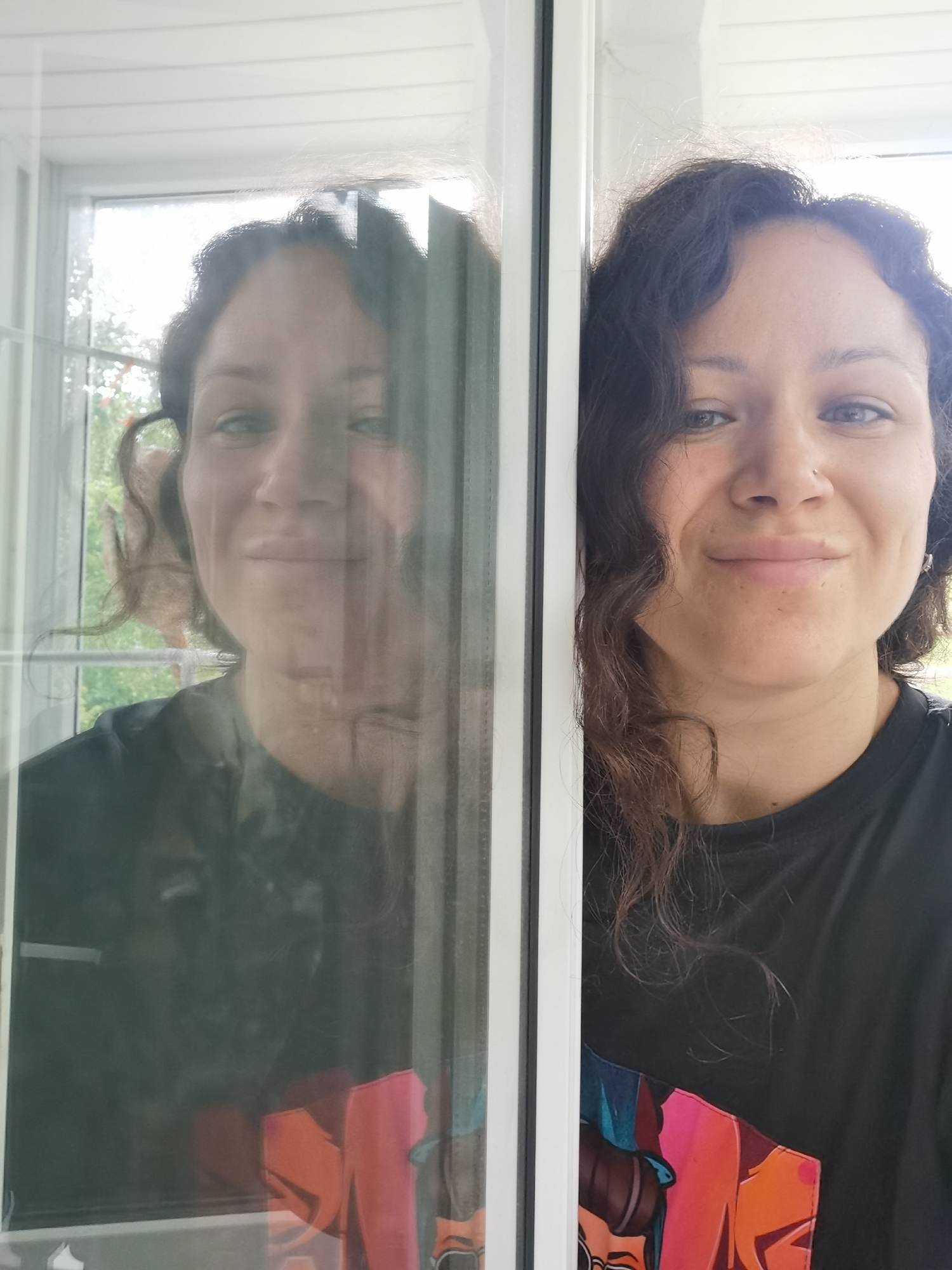 In the city of Chaikovsky, Russia, a woman with dark, curly hair and fair skin takes a selfie, her image captured partly through a glass surface, creating a reflection. She is wearing a black t-shirt with a colorful graphic design. Her left side is visible through the glass, showing her reflection which is slightly blurred and appears to be in an indoor setting with a window in the background. The woman's visible eye on the right side of the image is looking directly at the camera, and she has a slight smile. Her nose is slightly to the left, and her lips are closed. A subtle nose ring is visible on her right nostril. Her hair cascades over her shoulders and frames her face. The background on the right side of the image reveals a glimpse of an outdoor scene with green foliage. The glass surface, likely a window or door, has a white frame, dividing the woman's direct image from her reflection.