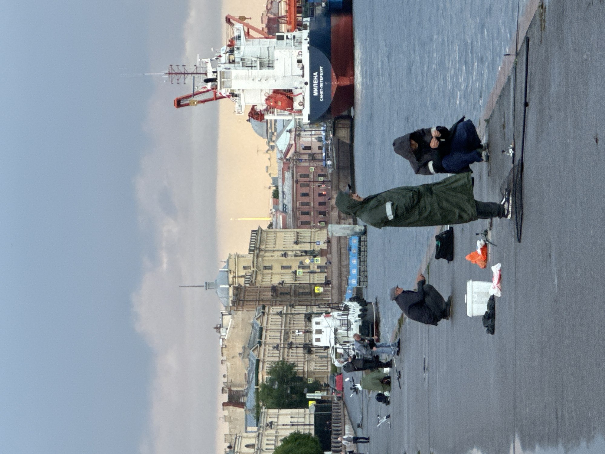 The image depicts a scene along the Neva River embankment in Saint Petersburg, Russia. The sky is overcast, with a mix of grey clouds and patches of pale yellow light. In the background, a large cargo ship, identified by the name "МИЛЕНА" and "САНКТ-ПЕТЕРБУРГ" on its hull, is docked. It is predominantly white and red, with a prominent crane structure.
Along the concrete embankment in the foreground, several individuals are engaged in fishing. One person, wearing a dark grey coat and a cap, is crouched down, seemingly holding a fishing rod. Another individual, dressed in a long green hooded coat, stands to the left. To the right of the crouching fisherman, another person, wearing a dark hooded jacket and blue pants, is also crouched down and appears to be preparing fishing tackle. In front of them are various fishing accessories, including rods, tackle boxes, and bags.
The embankment is wet, suggesting recent rain. Further back on the embankment, a small white boat is moored. The city's architecture lines the background, characterized by numerous classical buildings with ornate facades and mansard roofs, typical of Saint Petersburg. The overall atmosphere is subdued, likely due to the cloudy weather.