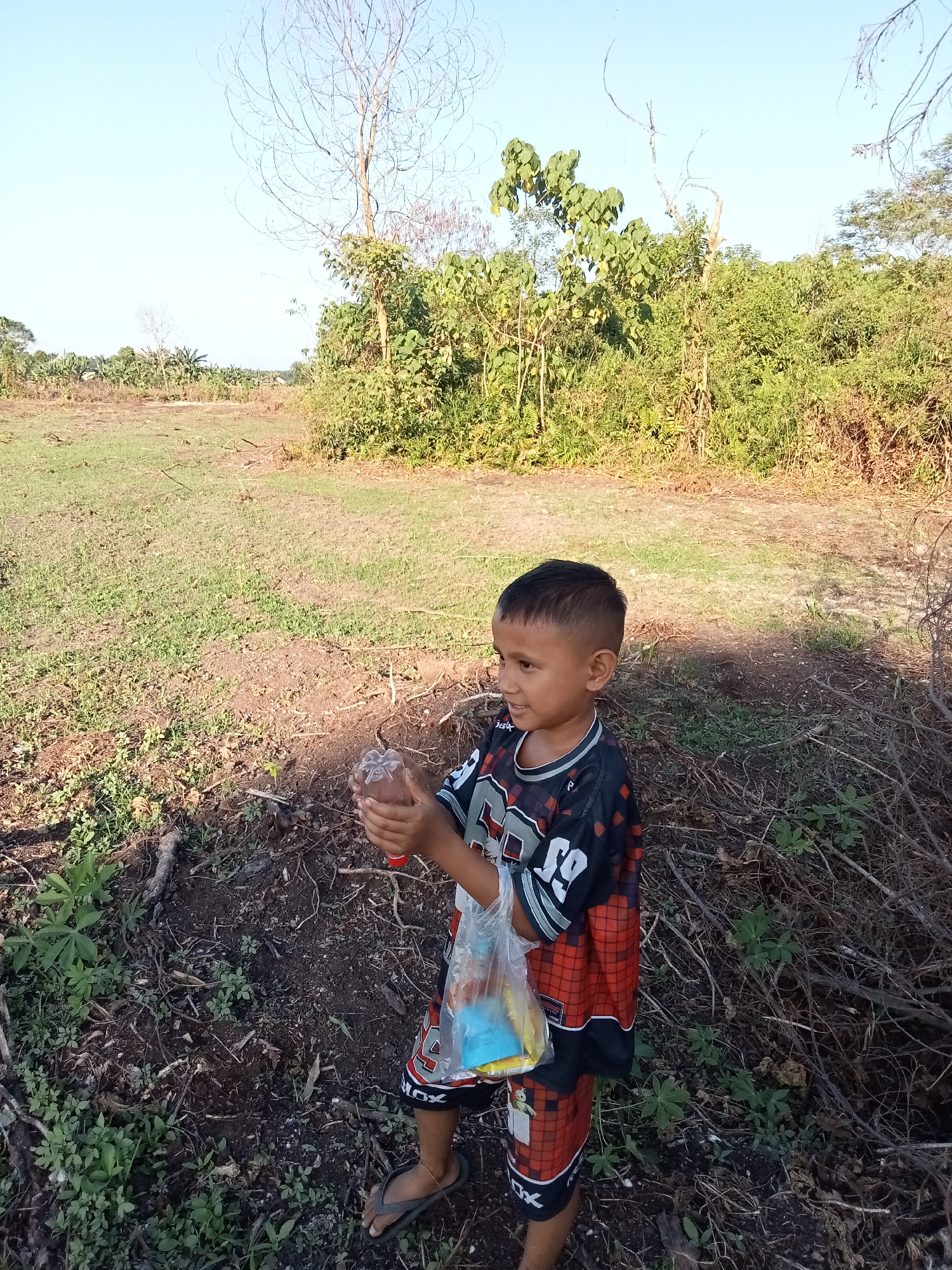 A young boy stands in a sunlit, dry field, holding a clear plastic bottle containing what appears to be candy. He is wearing a black and red checkered jersey with white numbering and black shorts with a red and white checkered pattern. The background consists of dry grass, scattered debris, and dense vegetation. In the distance, there are trees and a pale blue sky. The scene appears to be an outdoor, rural setting, possibly in Teluk Kenidai, Indonesia.