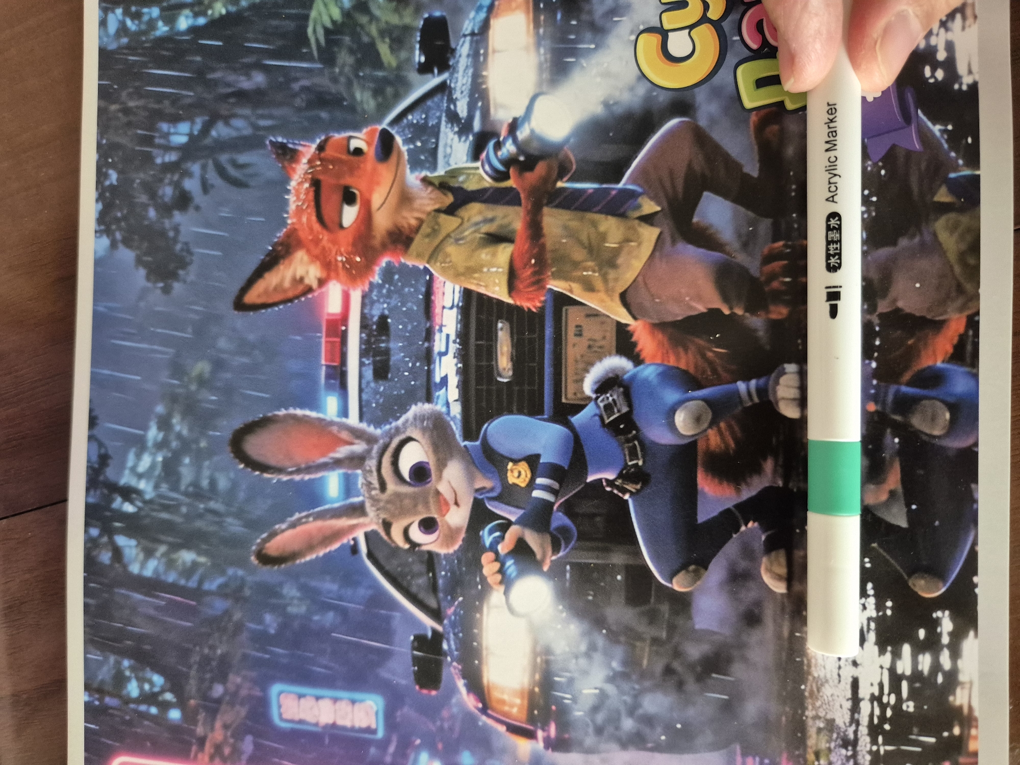 The image displays a poster featuring two animated anthropomorphic characters, a rabbit police officer named Judy Hopps and a fox police officer named Nick Wilde, from the movie Zootopia. They are standing in front of a police car during a rainstorm. The characters are positioned in a crouched stance, holding flashlights. The background depicts a city street at night with neon lights and wet surfaces reflecting the illumination. The lower part of the image shows a white marker with green accents and Chinese and English text indicating "Water-based Ink Acrylic Marker". The scene is not identifiable as being located in Kokshetau, Kazakhstan, or any specific city.
