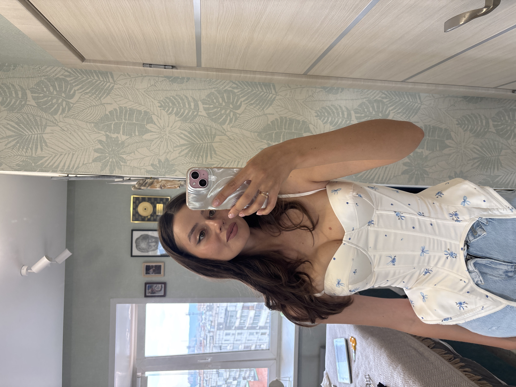 A woman is taking a selfie in a well-lit room. She is wearing a white corset with a blue floral pattern and blue denim jeans. The room has light green walls and a door with a wood grain finish. A window in the background offers a view of city buildings. The woman is holding a silver smartphone in her right hand, angled to capture her reflection in a mirror. On a table to her left, there's a rectangular object and a pair of orange-handled scissors. The woman's hair is dark brown and flows over her shoulders. She has makeup on and is wearing two rings on her right hand. The scene appears to be a domestic setting within an apartment in Krasnoyarsk, Russia, given the architectural style of the buildings visible through the window.