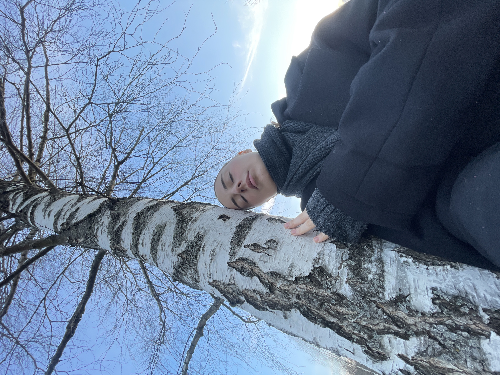 A low-angle shot depicts a person leaning against a birch tree. The person has a shaved head and is wearing a dark-colored coat and gray scarf. The birch tree has white bark with black markings. The sky is visible in the background, with a few thin clouds. Bare tree branches are visible extending upward and to the left. The location is Moscow, Russia.