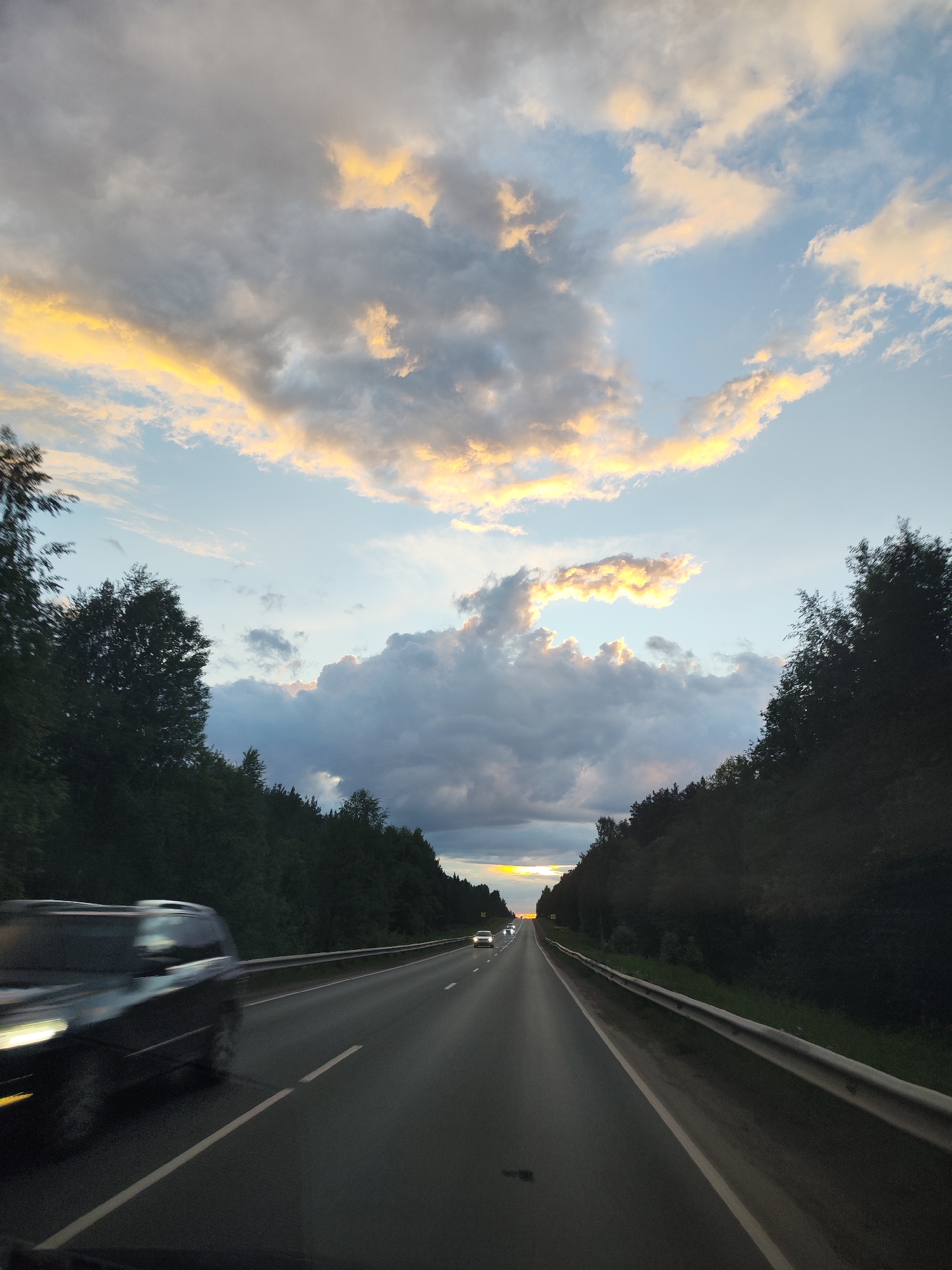 A view from inside a moving vehicle looking out onto a highway. The highway is flanked by dense green trees on both sides. The sky above is filled with clouds, some illuminated with a warm yellow hue, suggesting either sunrise or sunset. A car is partially visible in the left foreground, blurred from motion. In the distance, headlights from another vehicle can be seen. The scene has a dynamic, fleeting quality due to the motion of the vehicle. The location can not be determined in the city of Chaikovsky, Russia, without additional information.